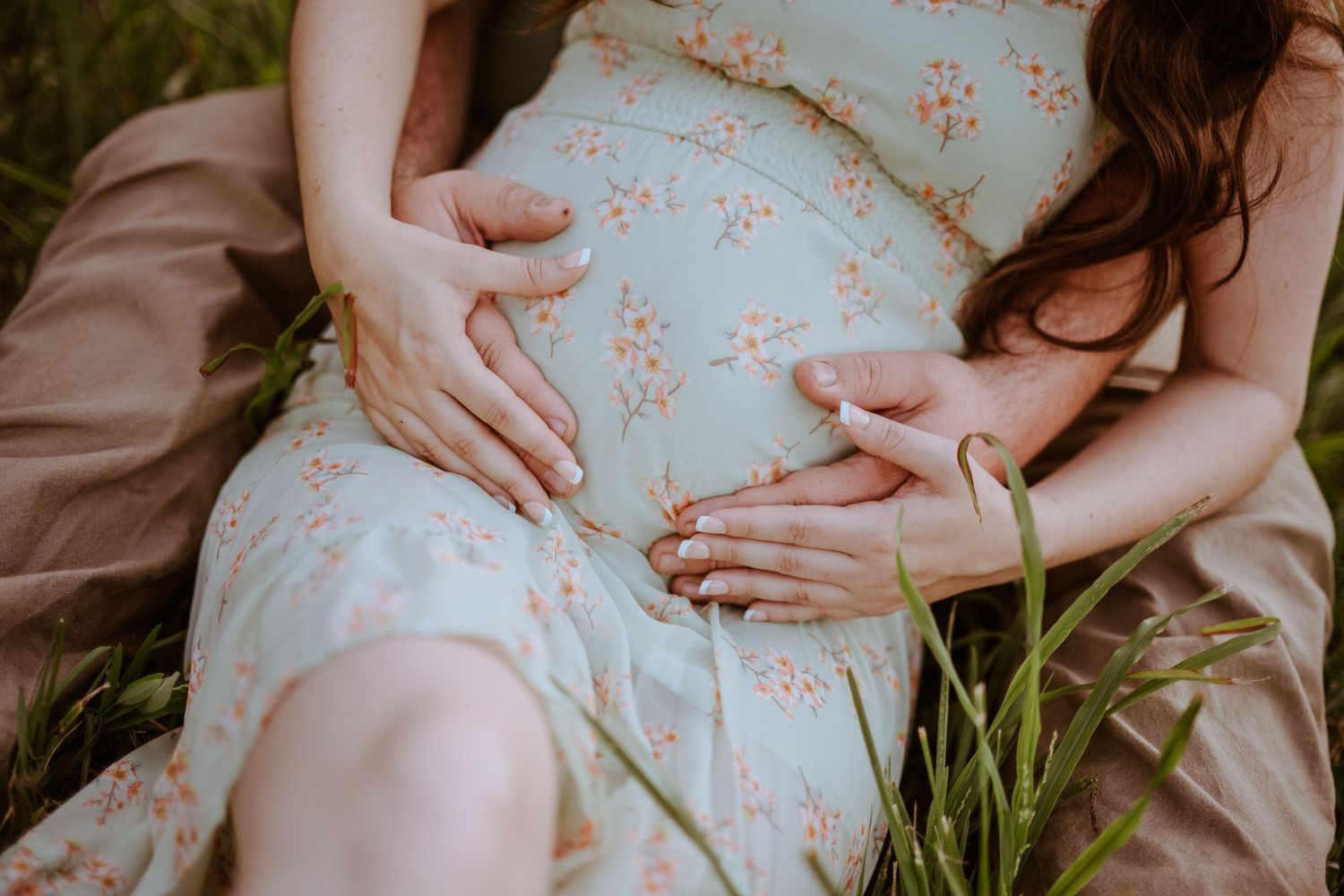 Close-up view of hands resting on a pregnant belly wearing a floral blue maternity dress while sitting in grass. Vanceburg Ky
