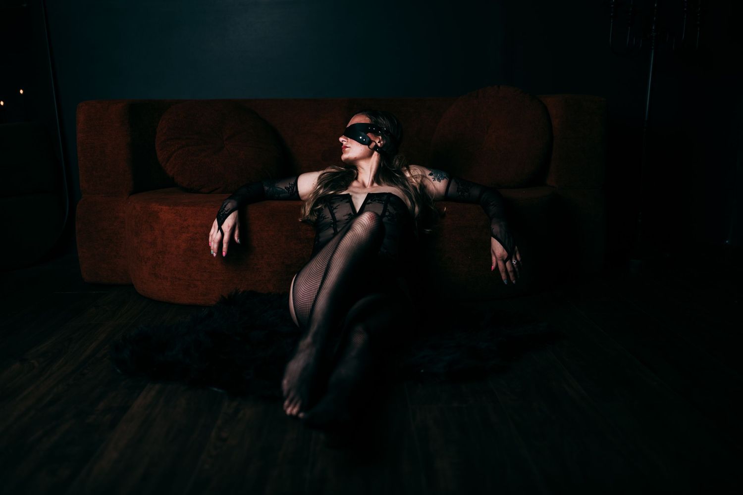 Woman in black lace bodysuit and blindfold poses dramatically on floor near a dark velvet sofa.