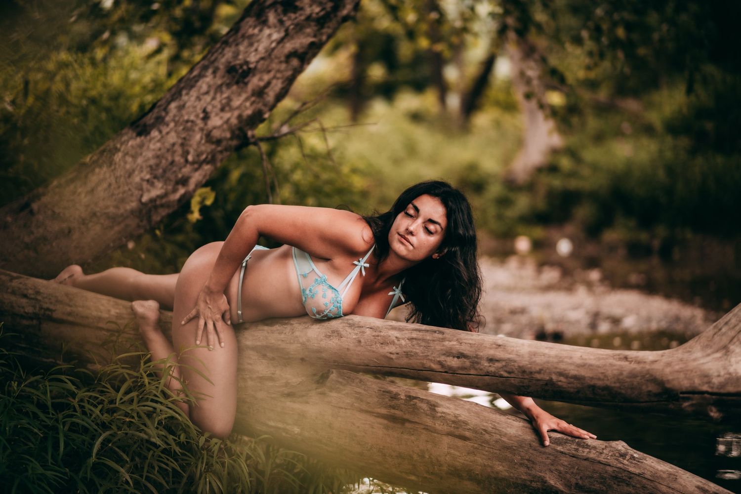 Woman in bikini posing gracefully on fallen logs near water surrounded by lush green trees.