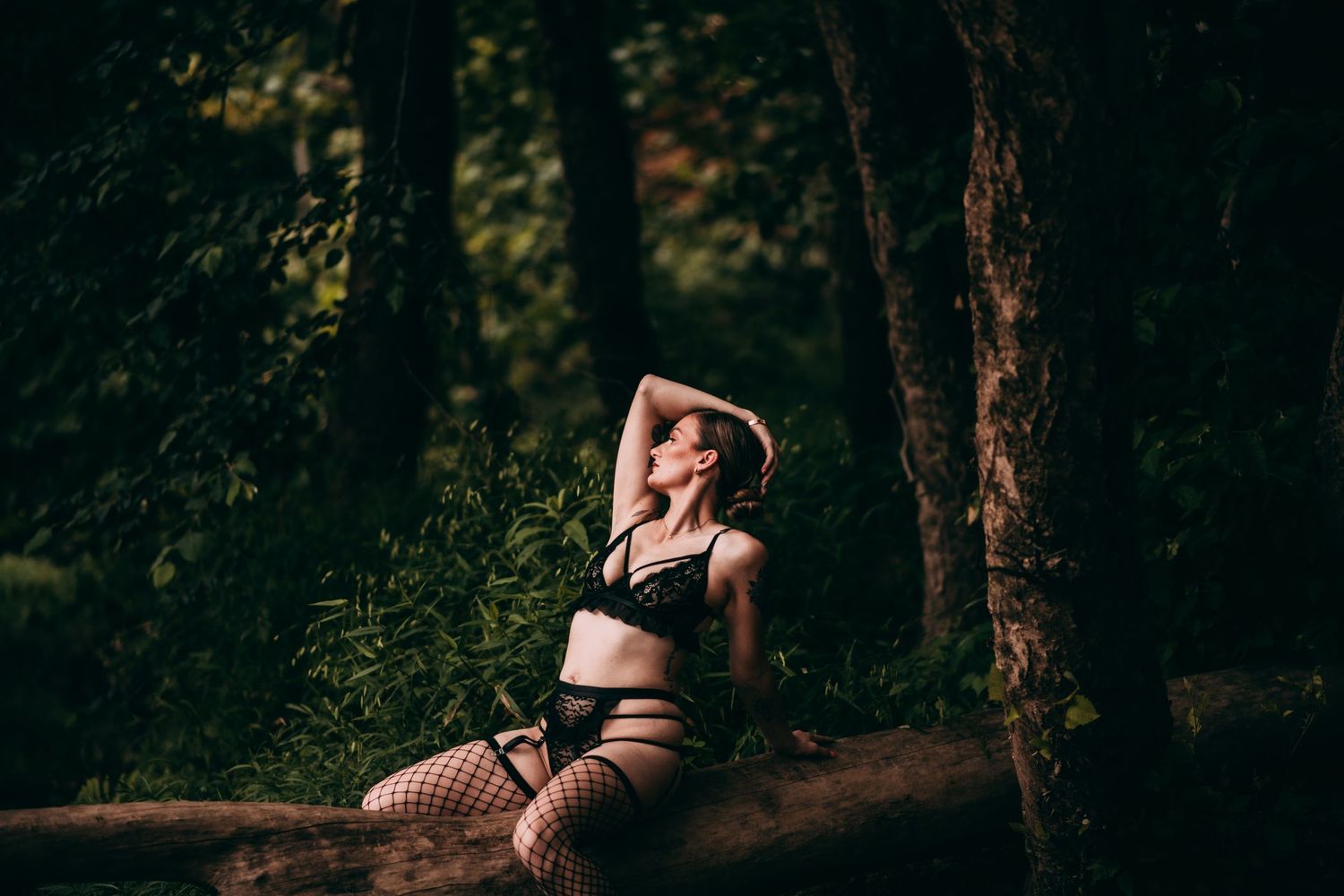 Woman in black lingerie and fishnet stockings posing on a log in a lush green forest setting.