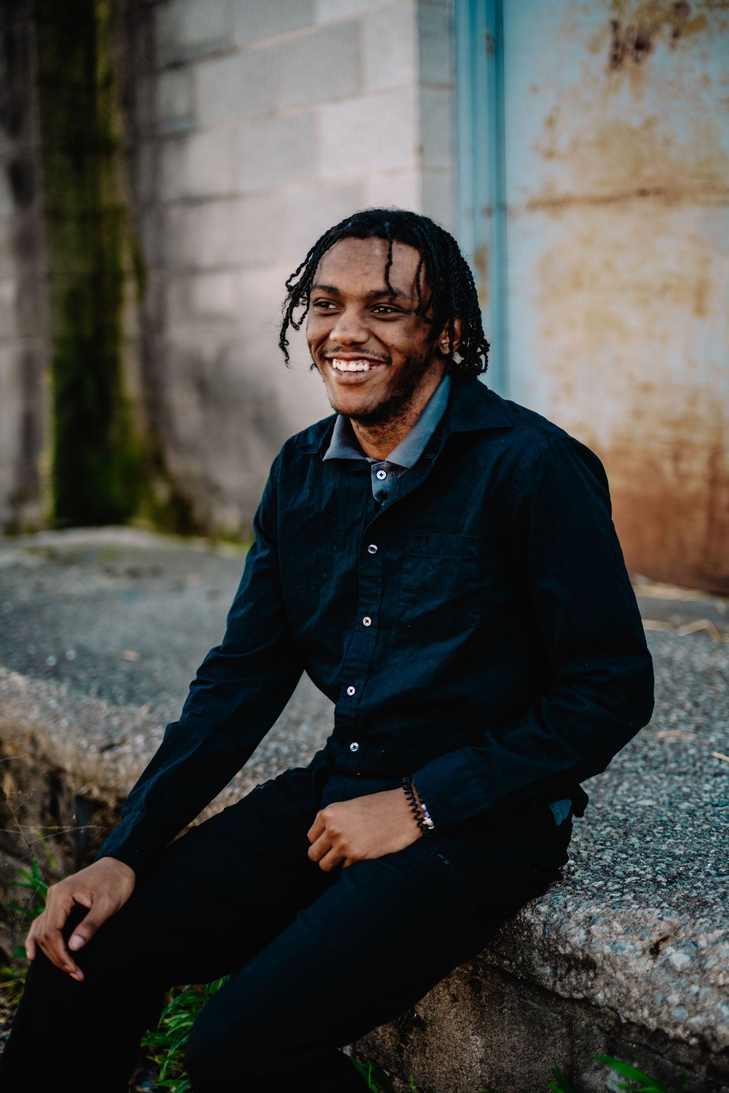Person in dark jacket smiling while sitting on concrete ledge outdoors.