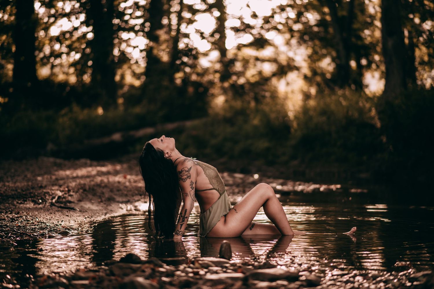 Woman with long dark hair posing in shallow forest stream at golden hour in artistic nature photo.