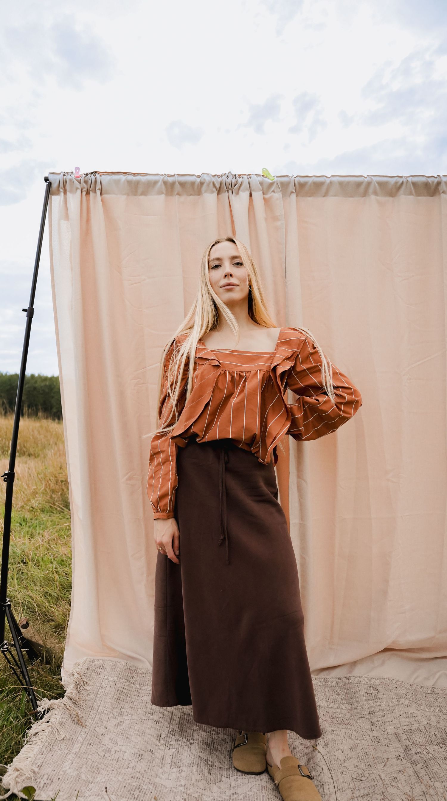 Blonde woman in rust striped top and dark brown maxi skirt standing before a light pink curtain backdrop outdoors.