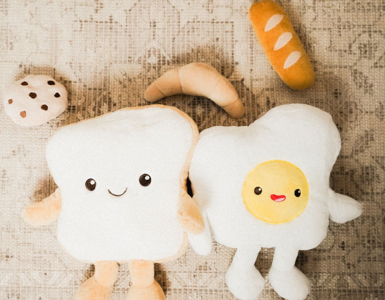 Cute bread slice and fried egg plush toys with smiling faces on a rug, with cookie and baguette plushies nearby.