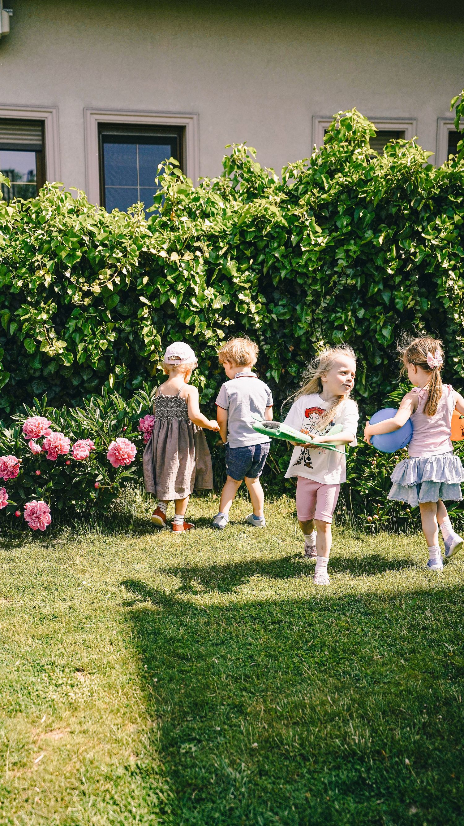 Four young children playing together in a sunny garden with pink hydrangeas and lush green hedges.
