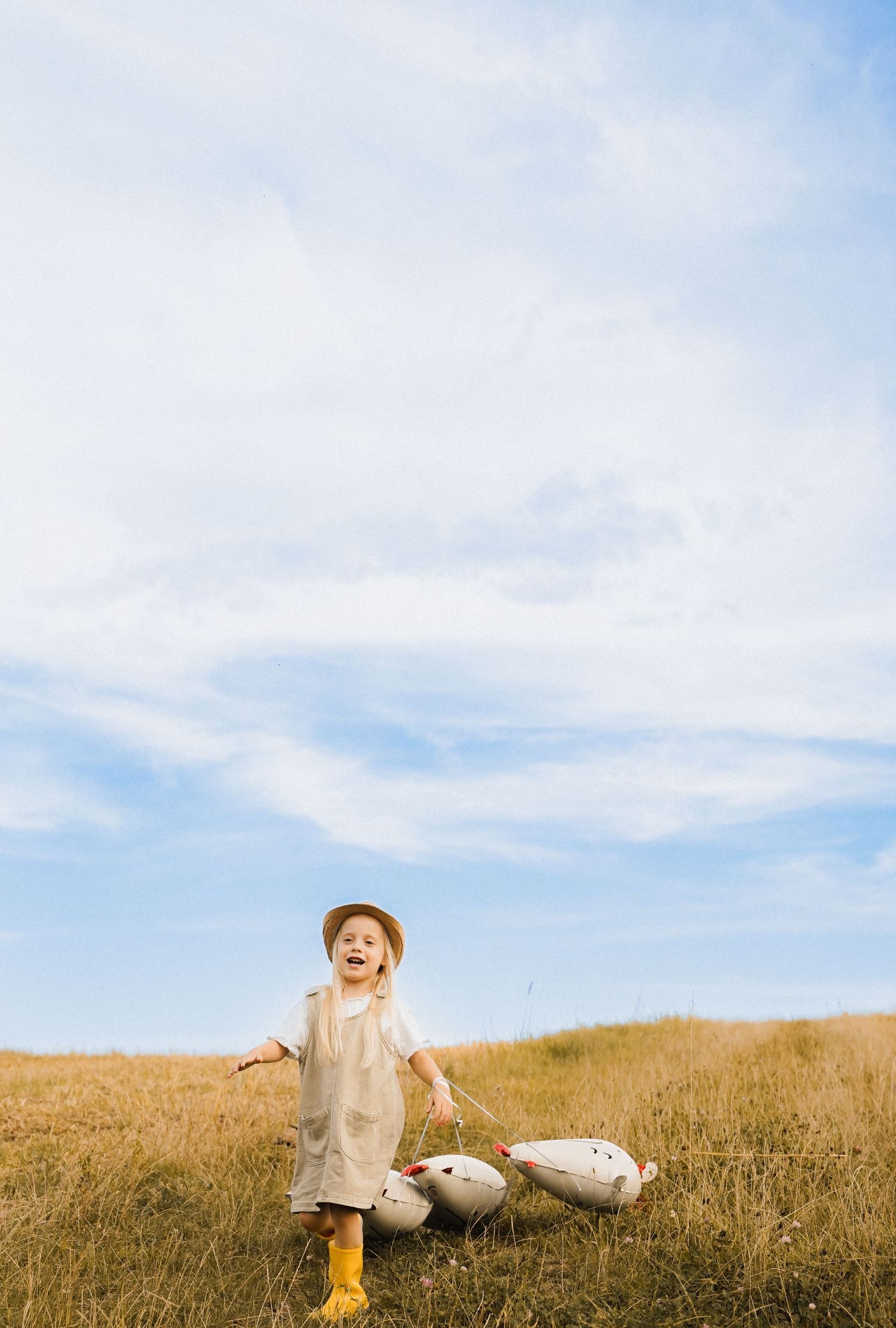 Joyful child in tan outfit and yellow boots laughing in golden field with toy geese under blue sky.