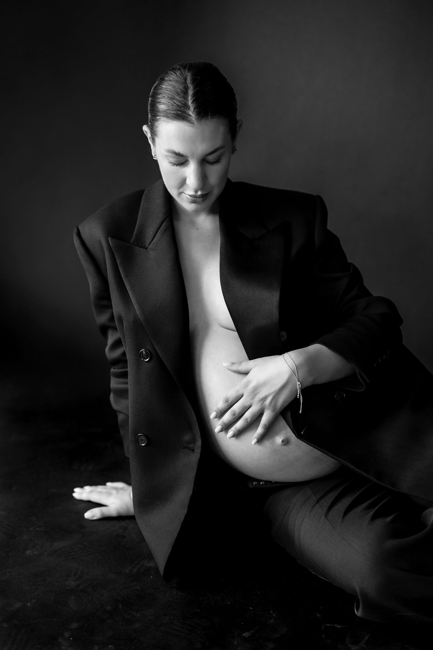 Black and white photo of a pregnant woman in an open blazer cradling her baby bump while seated on a dark floor.