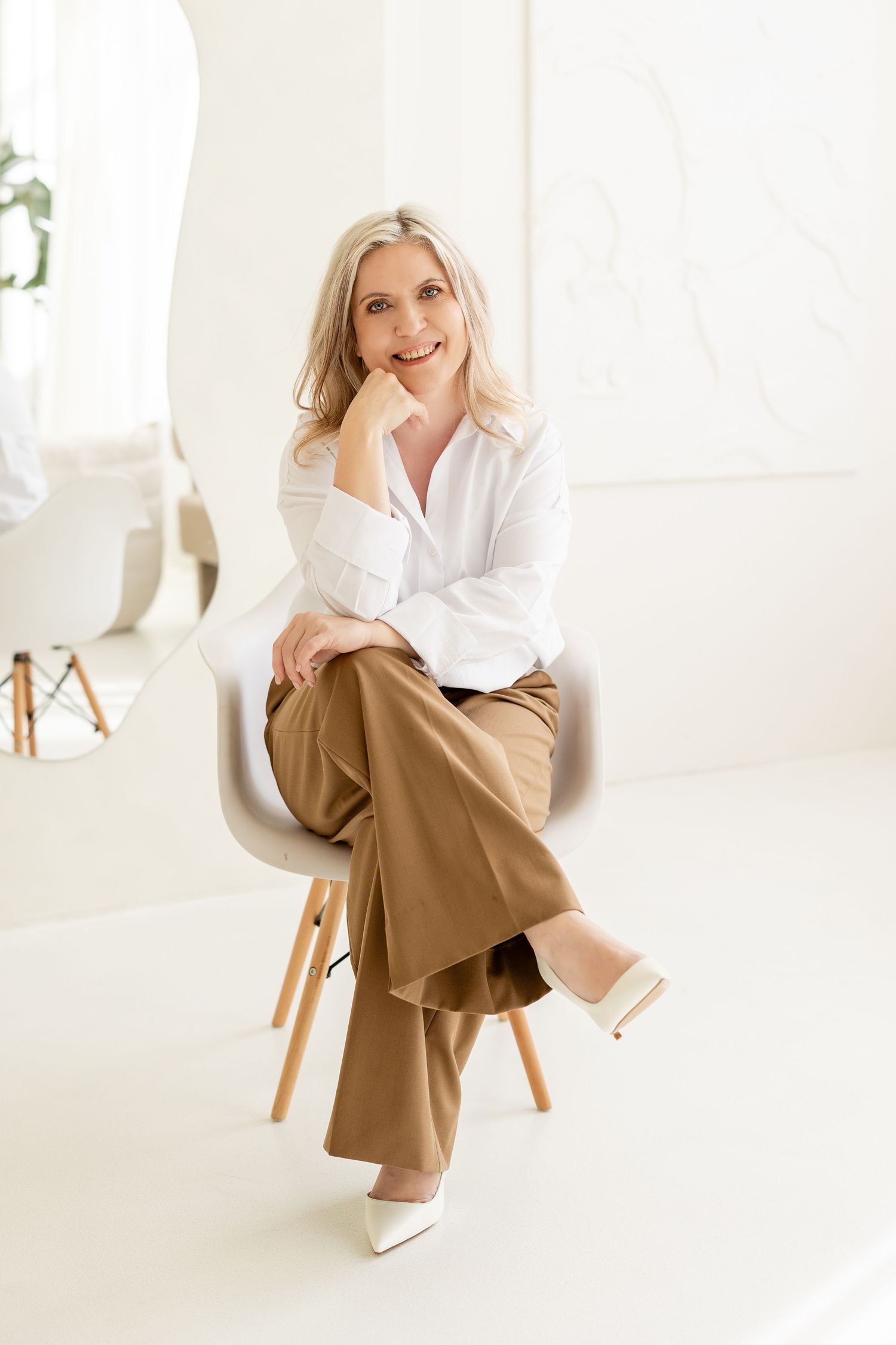 Professional portrait in bright white studio with modern chair, wearing crisp white shirt and tan wide-leg pants.