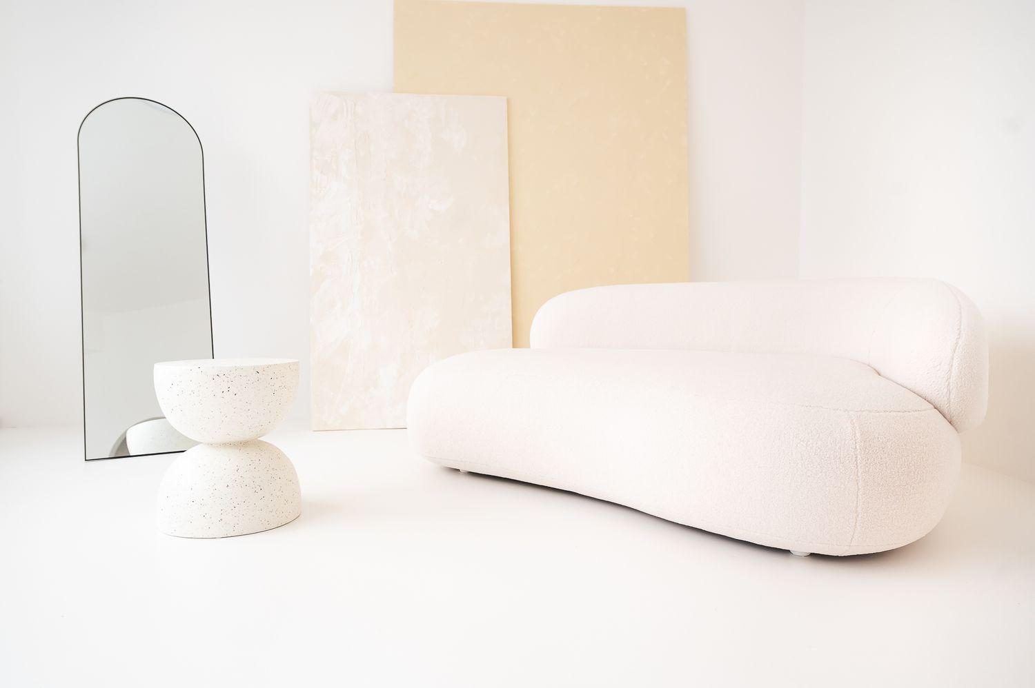 Minimalist white living room featuring a curved ivory sofa, sculptural bowl side table, arched mirror, and abstract art panels.