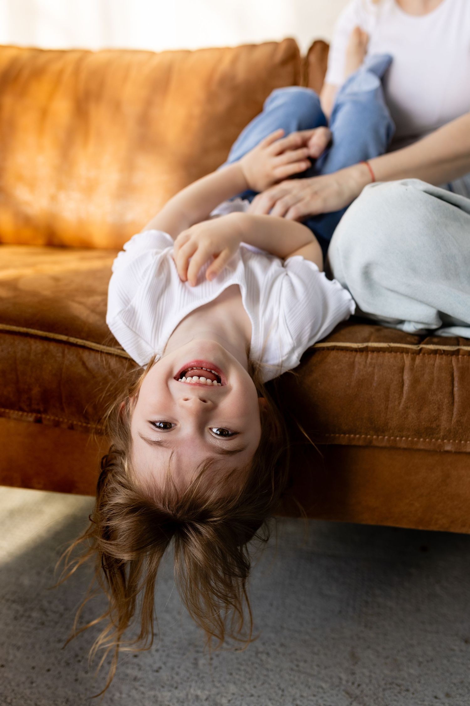 A playful child hangs upside down from a brown leather couch while laughing and having fun.