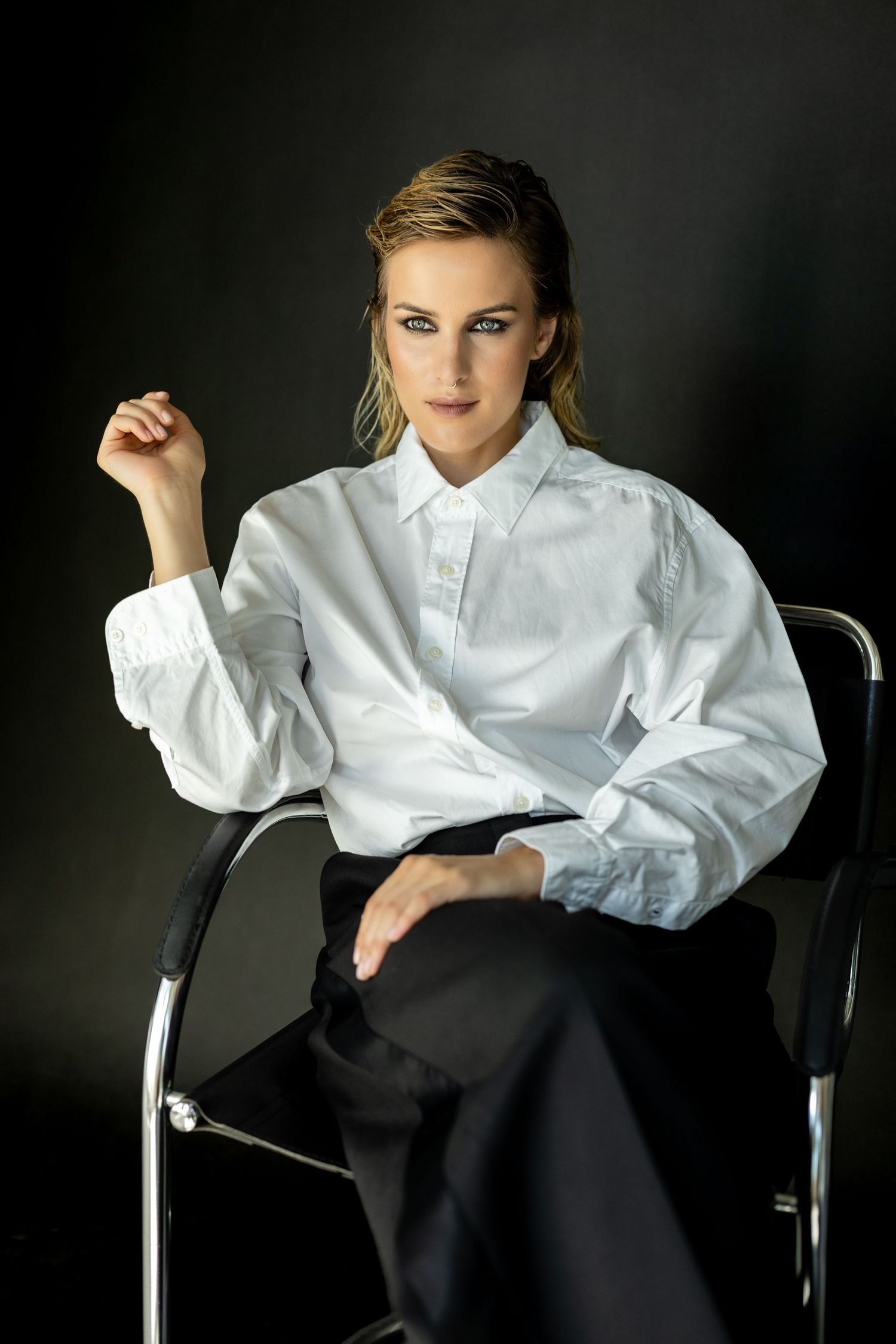 Woman in white oversized shirt and black trousers seated on chrome chair against dark background.