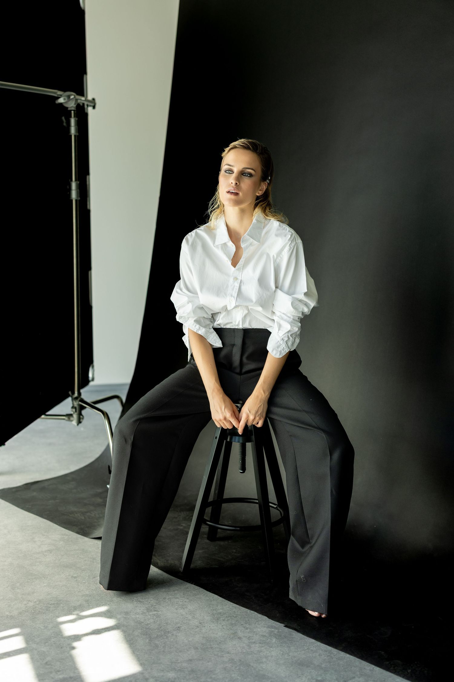 Woman in white oversized shirt and black wide-leg trousers sits on stool against dark backdrop in professional photo studio.