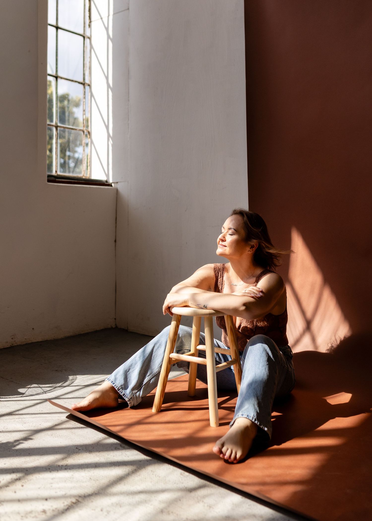 Woman sitting barefoot on floor leaning on wooden stool in sunlit room with warm shadows and jeans.
