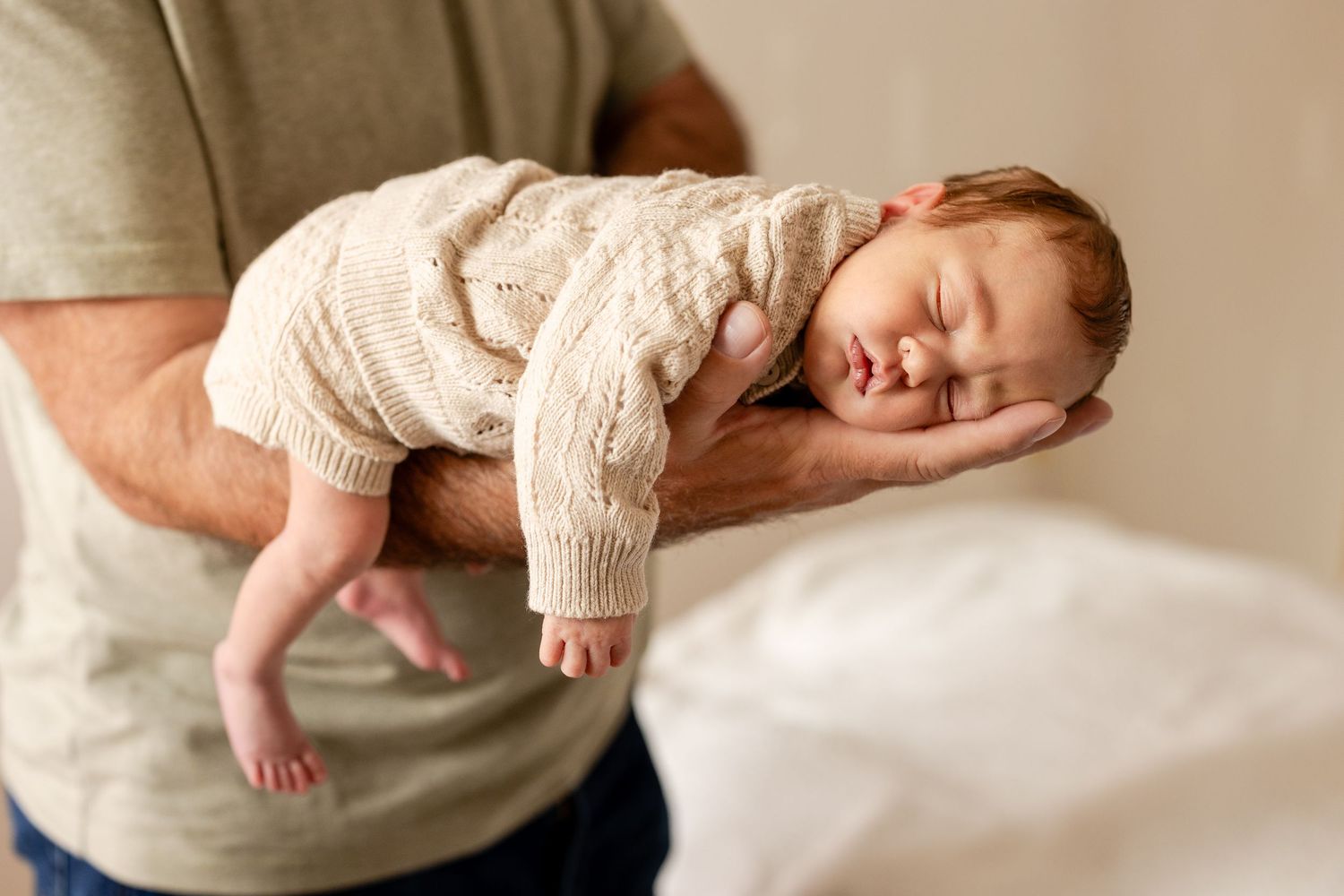 A father cradles a sleeping newborn baby in a knit onesie in his arms during a professional newborn photo session.