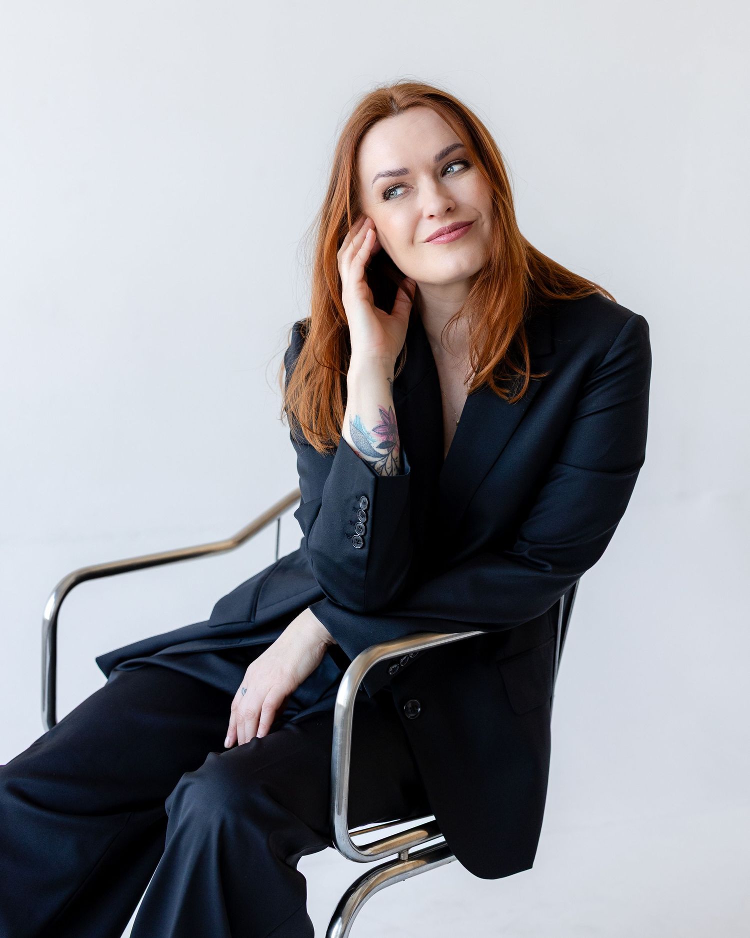 A woman with red hair in a black suit sits gracefully on a chrome chair against a white background.