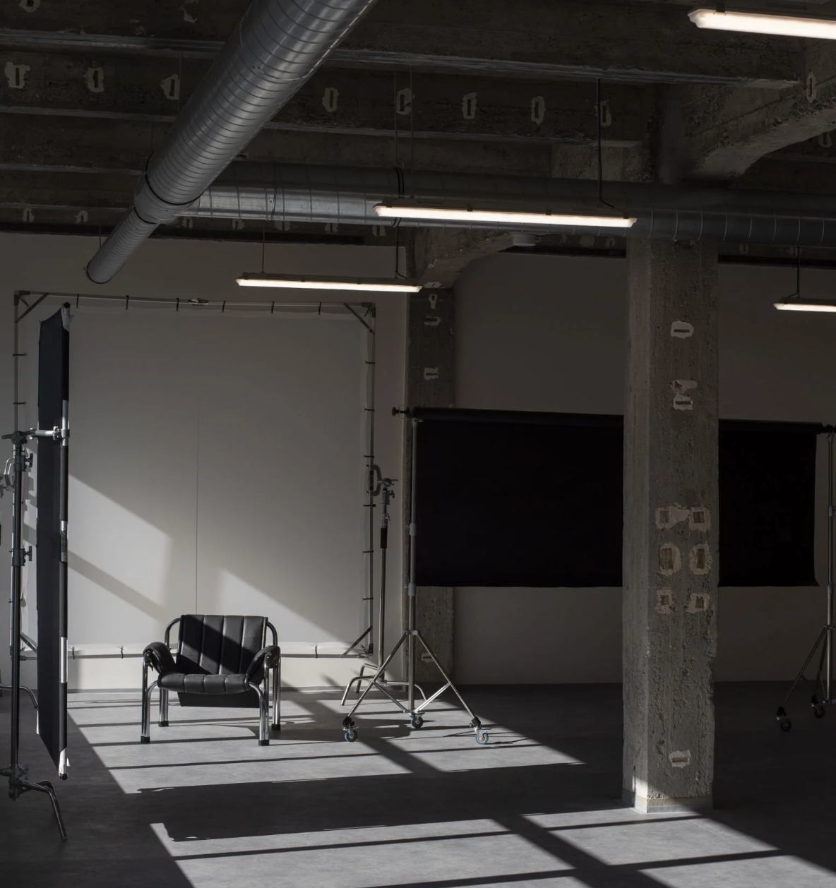 Empty photography studio with chair, backdrop, lighting equipment, and industrial ceiling pipes casting shadows on concrete floor.