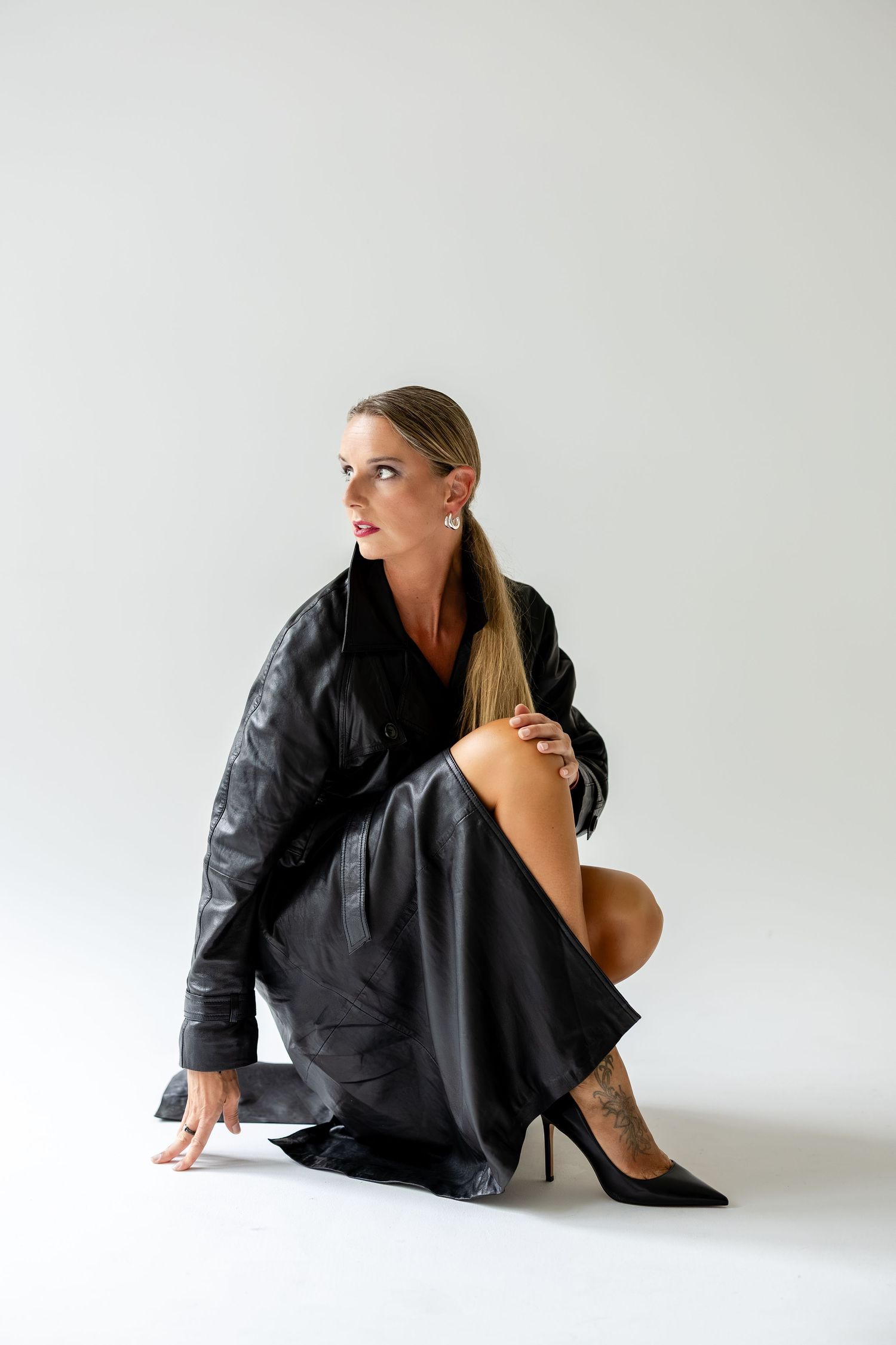 Woman in black leather jacket and heels crouching on white background, looking up with elegant pose.