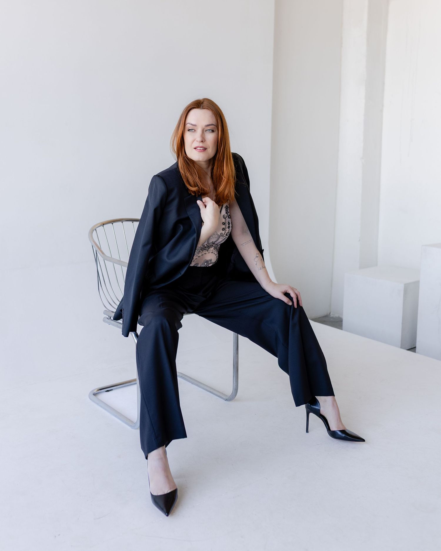 A person in a black business suit and heels sits on a modern wire chair against a white minimalist background.