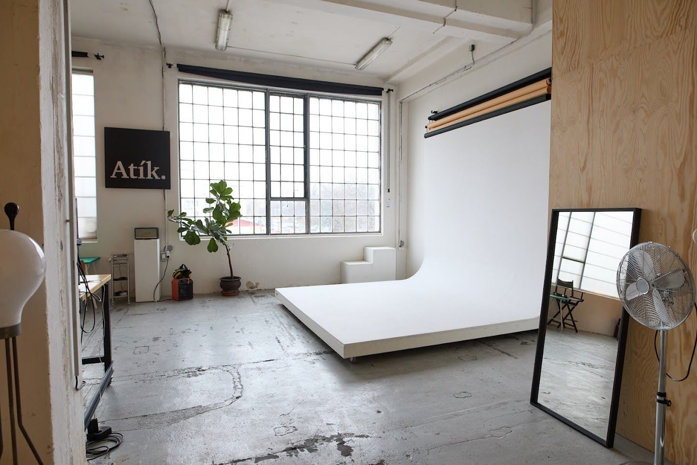 Industrial photography studio with white backdrop, concrete floors, large windows, mirror, and fan at Atik Studios.