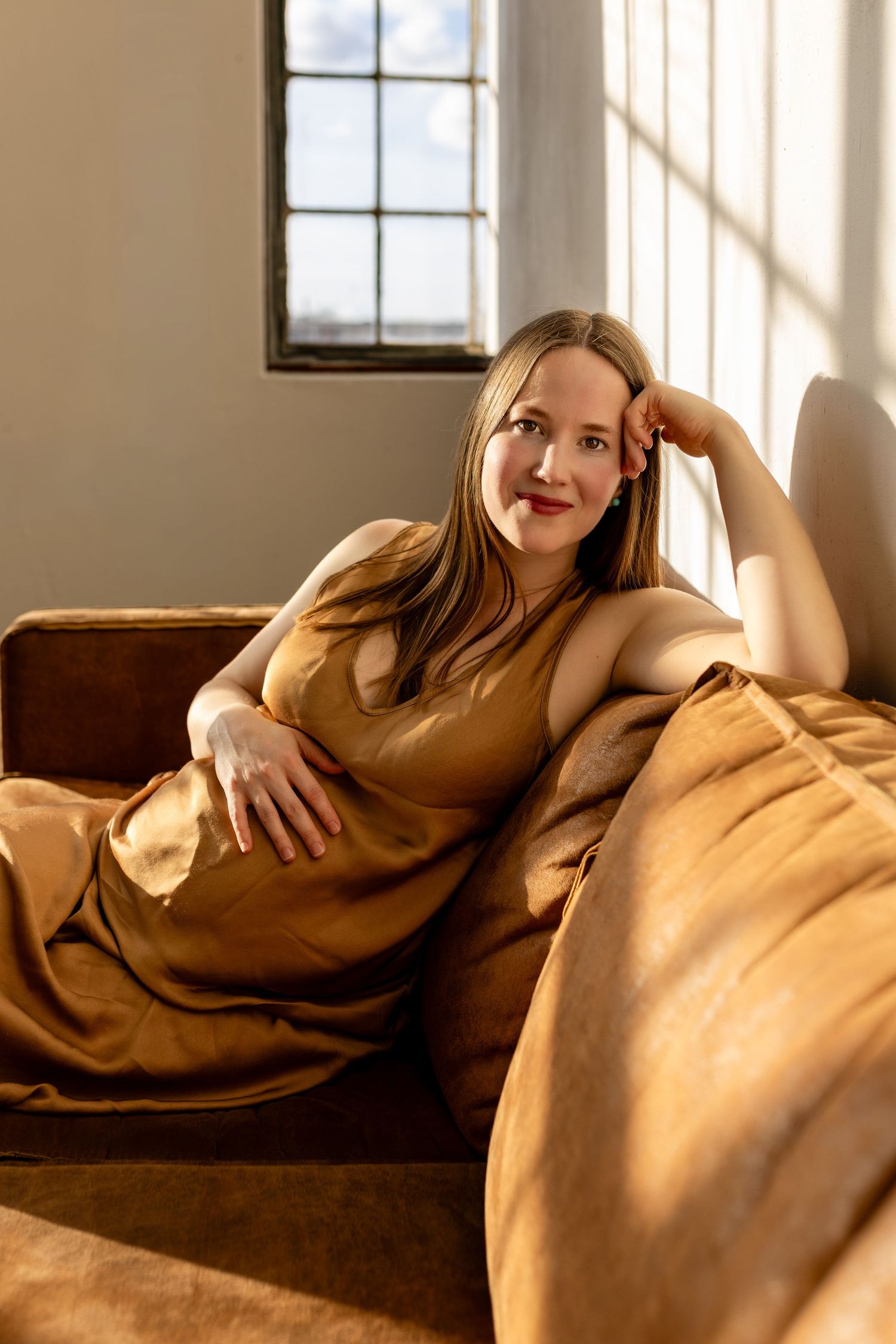 Person in a caramel-colored dress relaxes on a brown couch near a window with warm sunlight streaming in.