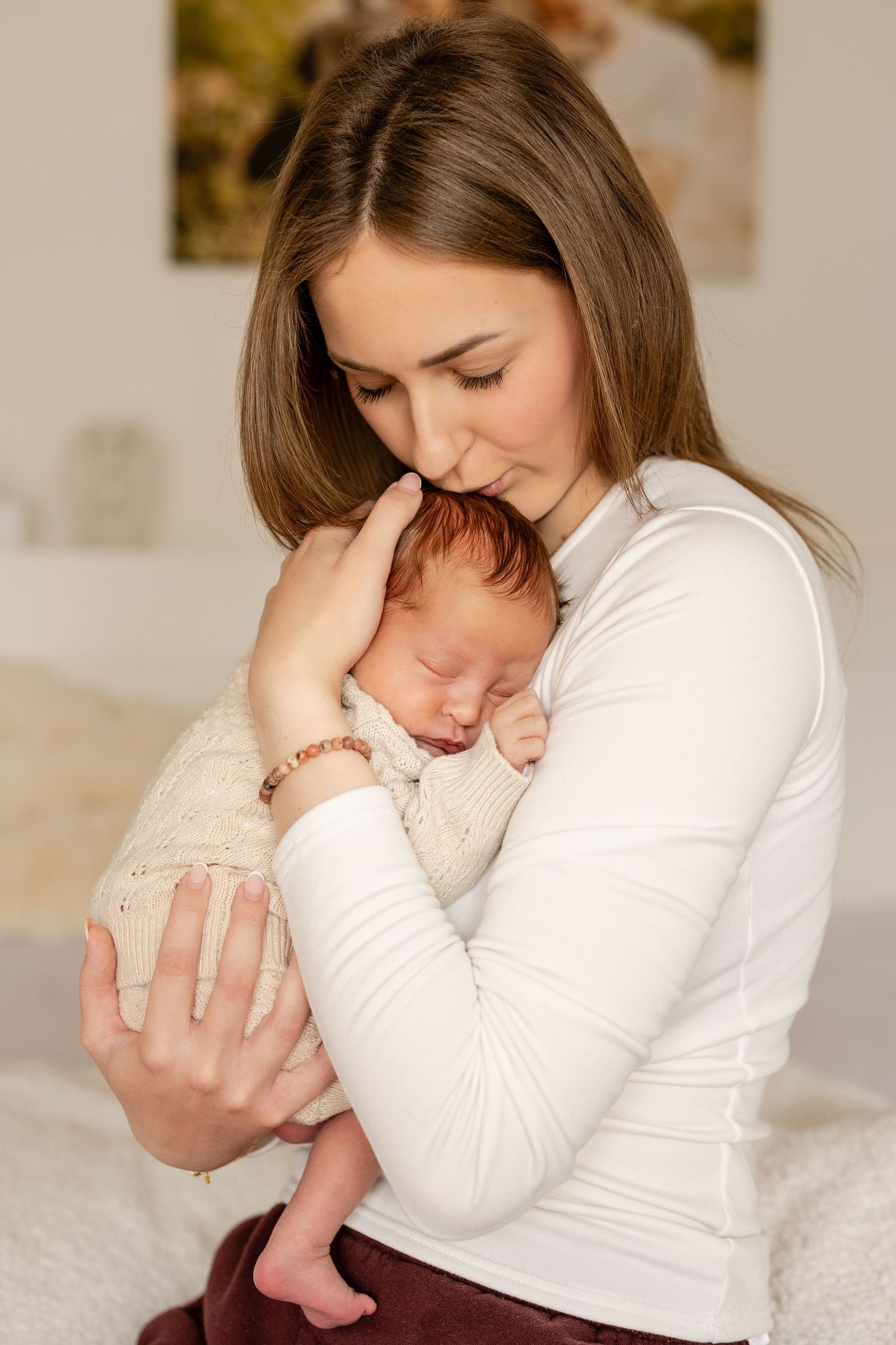 A loving mother in a white top gently kisses her sleeping newborn baby wrapped in a cream knit blanket.