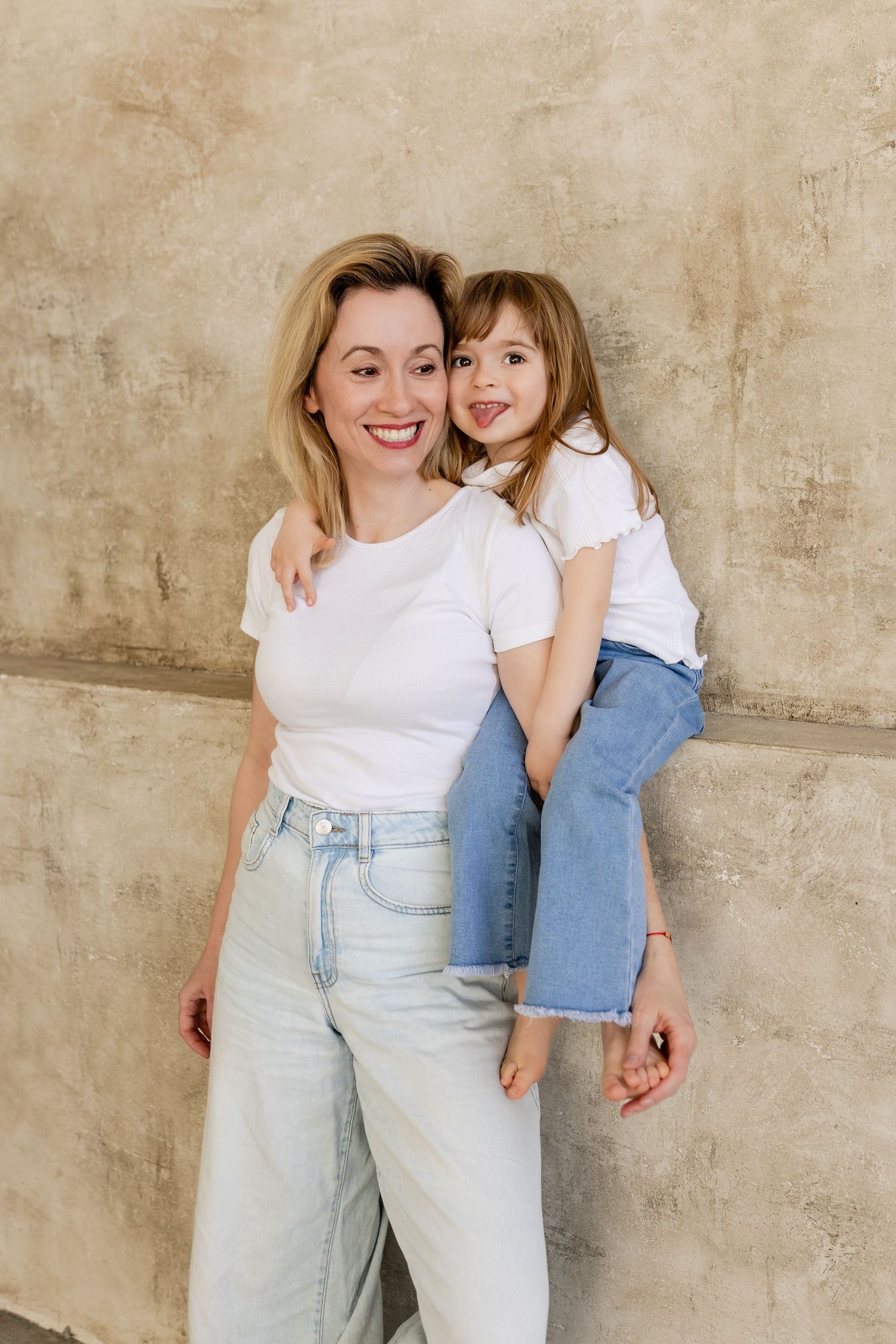 A parent and child wearing matching white shirts and light denim pose against a textured beige wall in a photo series.