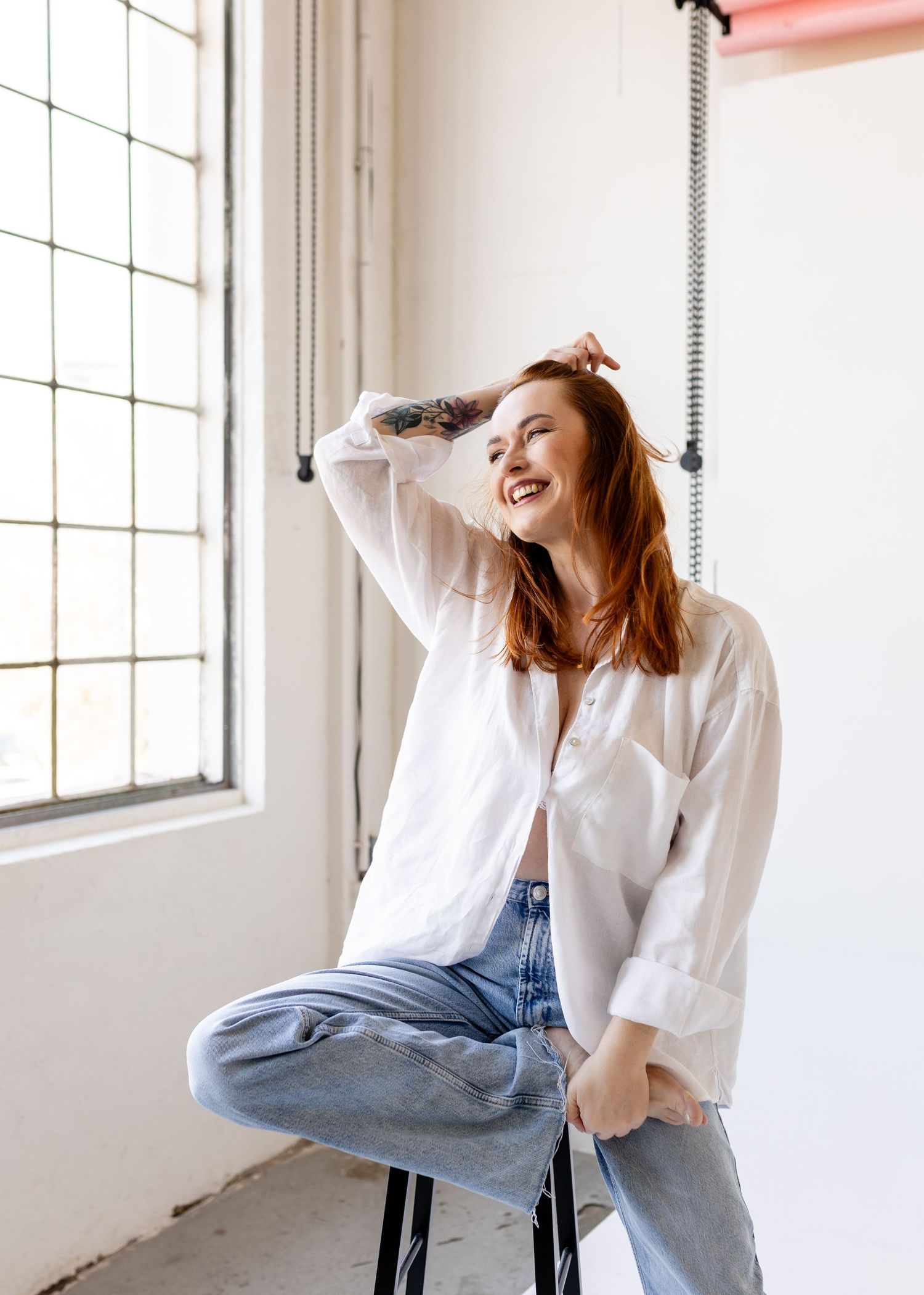 Smiling redhead woman in white shirt and jeans sitting on stool by window in bright studio.