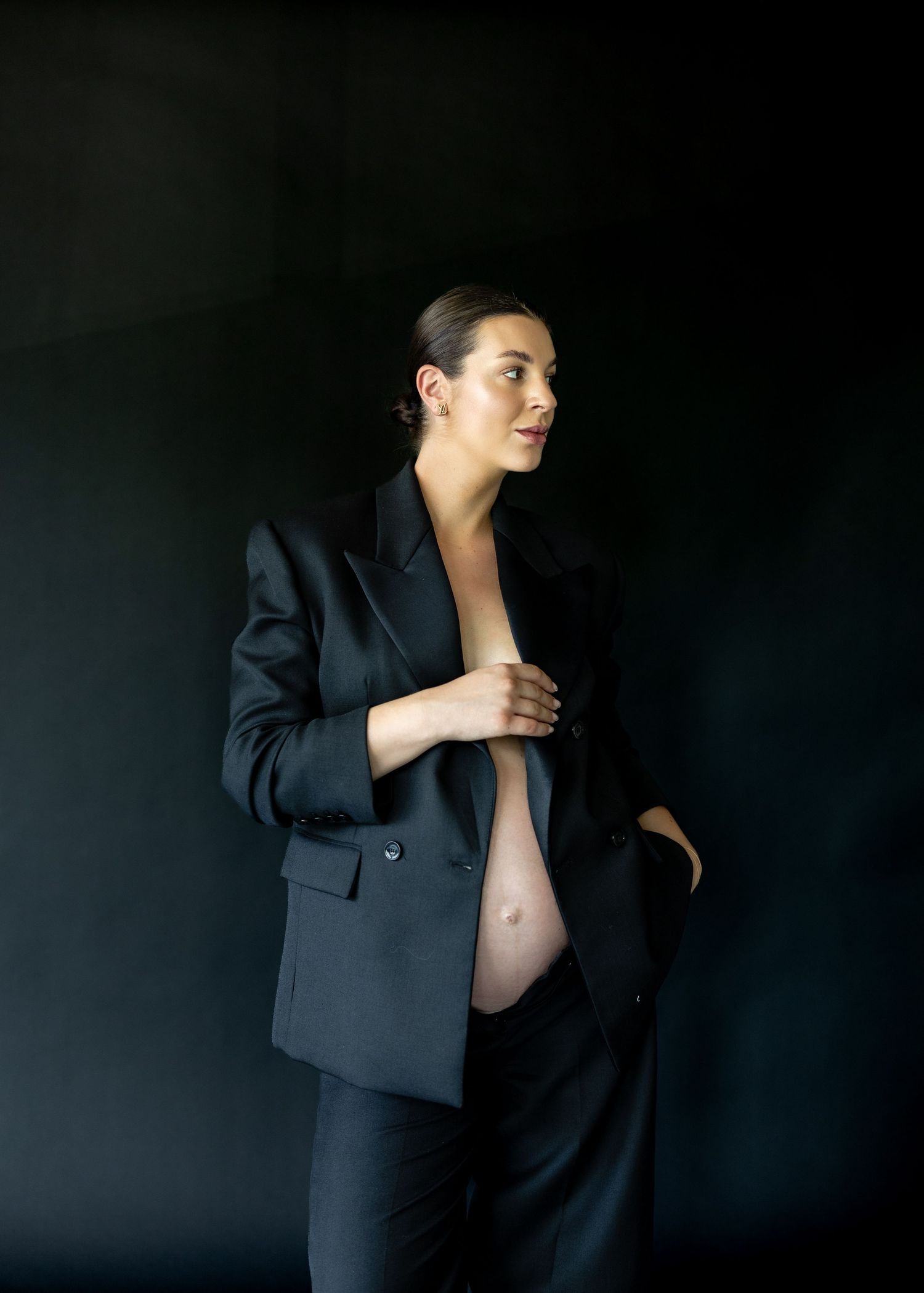 Pregnant woman in black blazer suit posing against dark background, showcasing maternity fashion with elegant style.