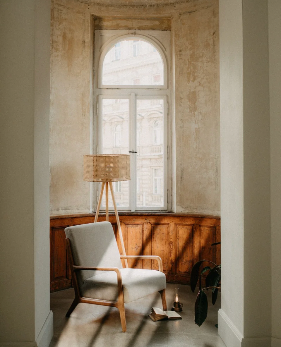 A cozy reading nook with a mid-century armchair, tripod floor lamp, and arched window in a rustic vintage room.