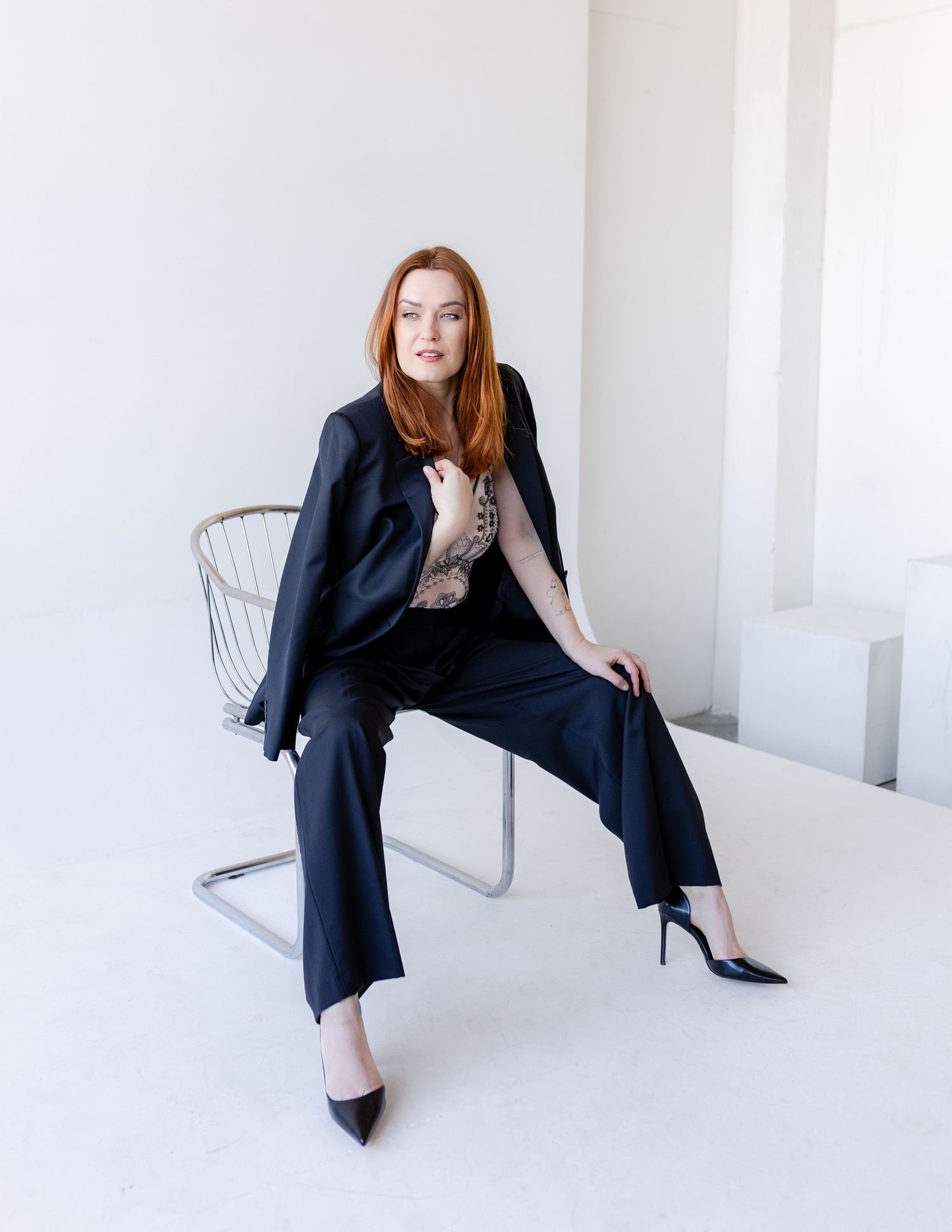 A redhead woman in a black suit and heels sits confidently on a metal chair in a bright white minimalist studio.