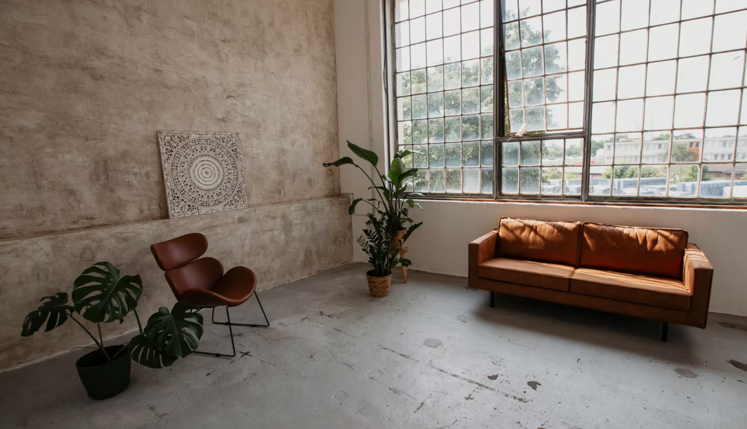 Modern industrial loft living room with brown leather sofa, monstera plants, and large windows on concrete floors.