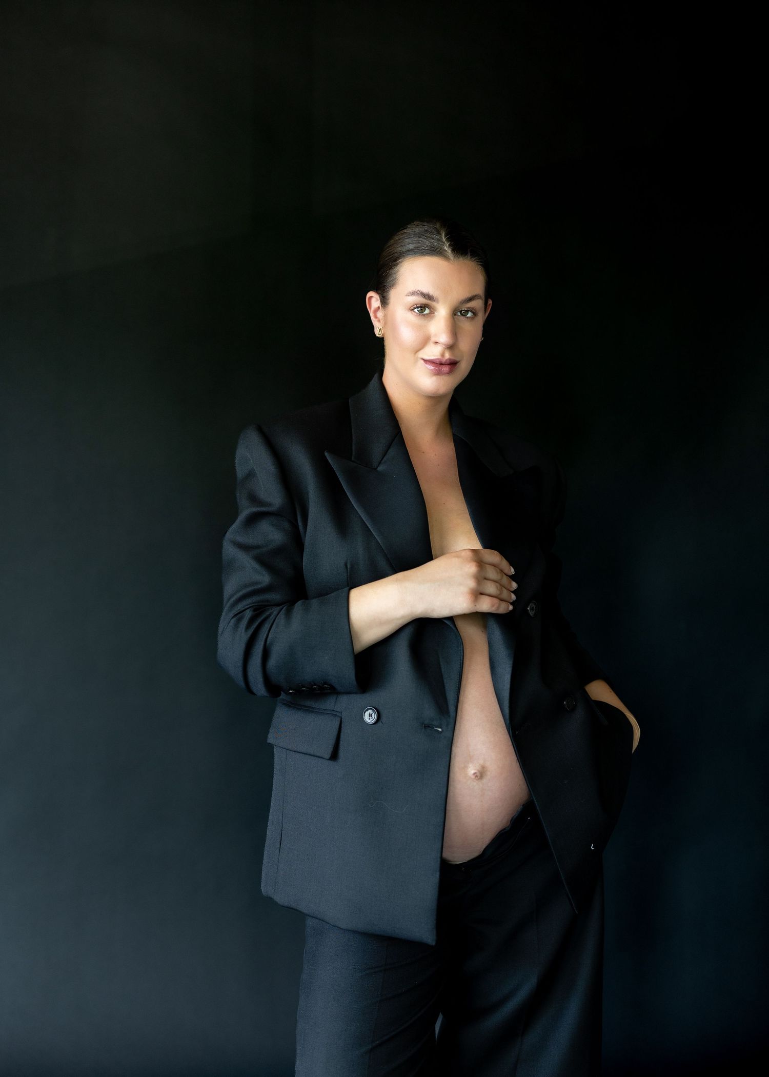 Pregnant woman in black blazer suit posing against dark background, showcasing baby bump in elegant maternity photo.