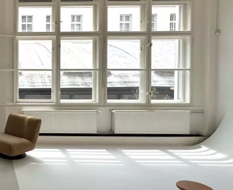 Minimalist white studio space with large windows, a brown accent chair, and a radiator casting natural light on white floors.