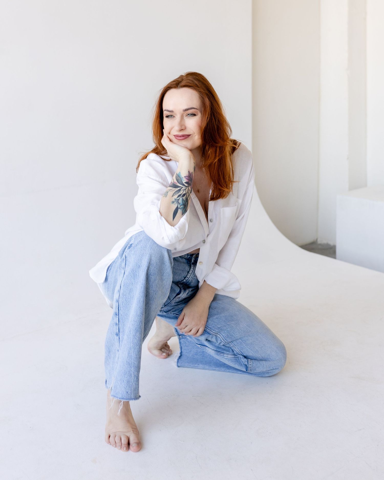 Redhead woman in white button-up shirt and light blue jeans kneeling barefoot on white floor.