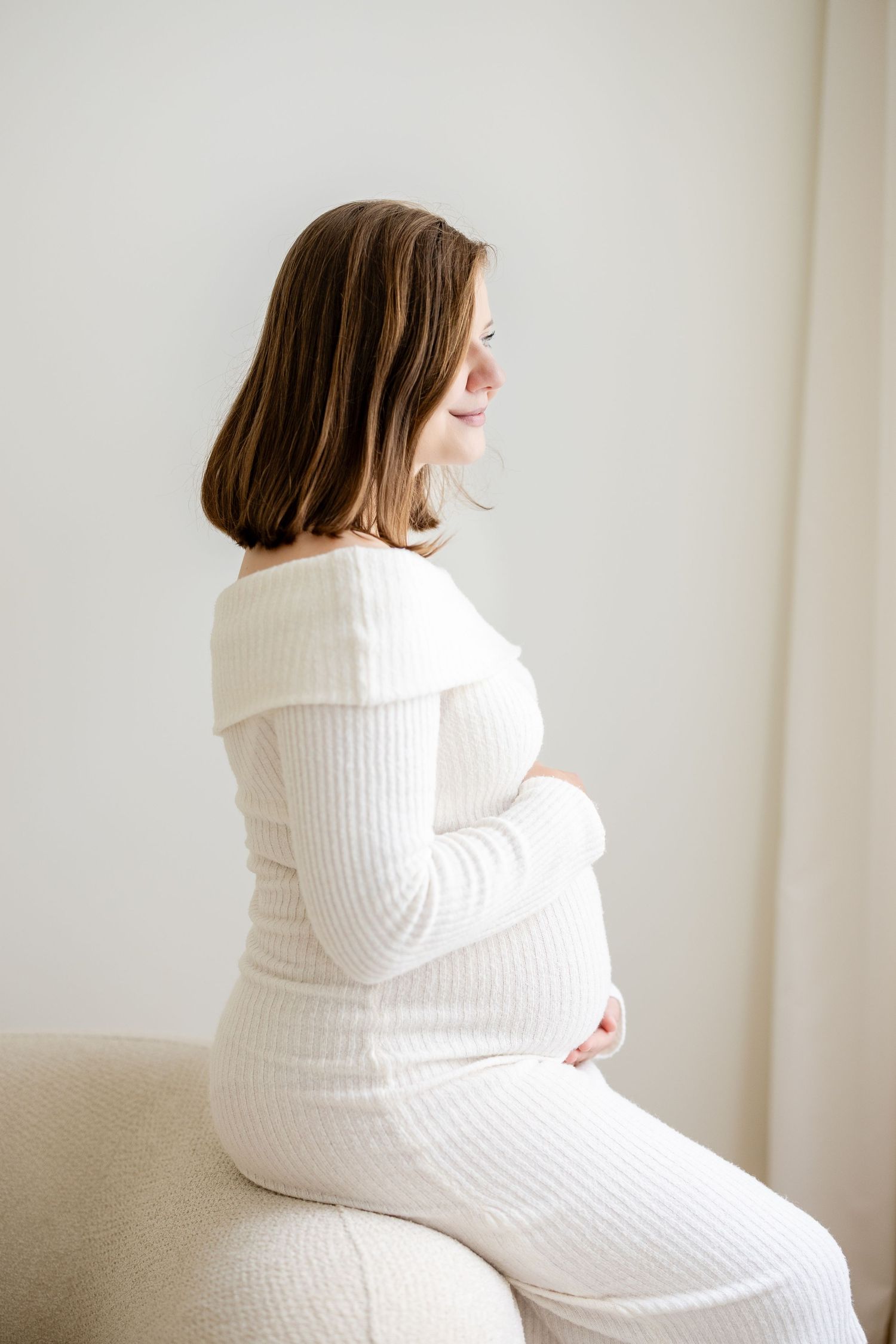 Smiling pregnant woman in white ribbed dress sitting sideways, cradling her baby bump against a light background.