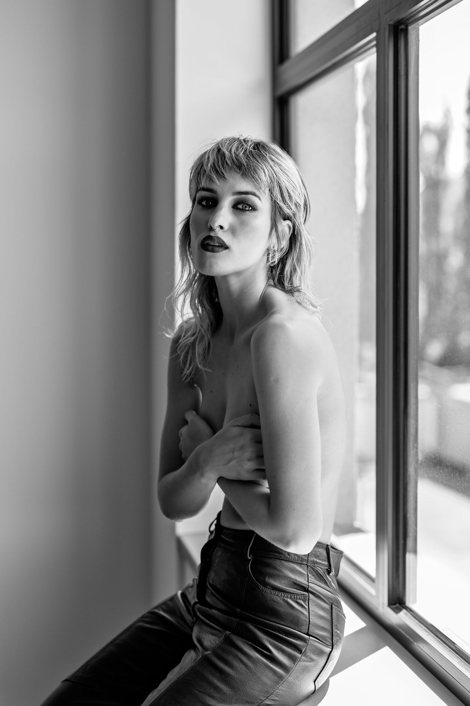 Black and white photo of a young woman with short blonde hair posing by a window in jeans.