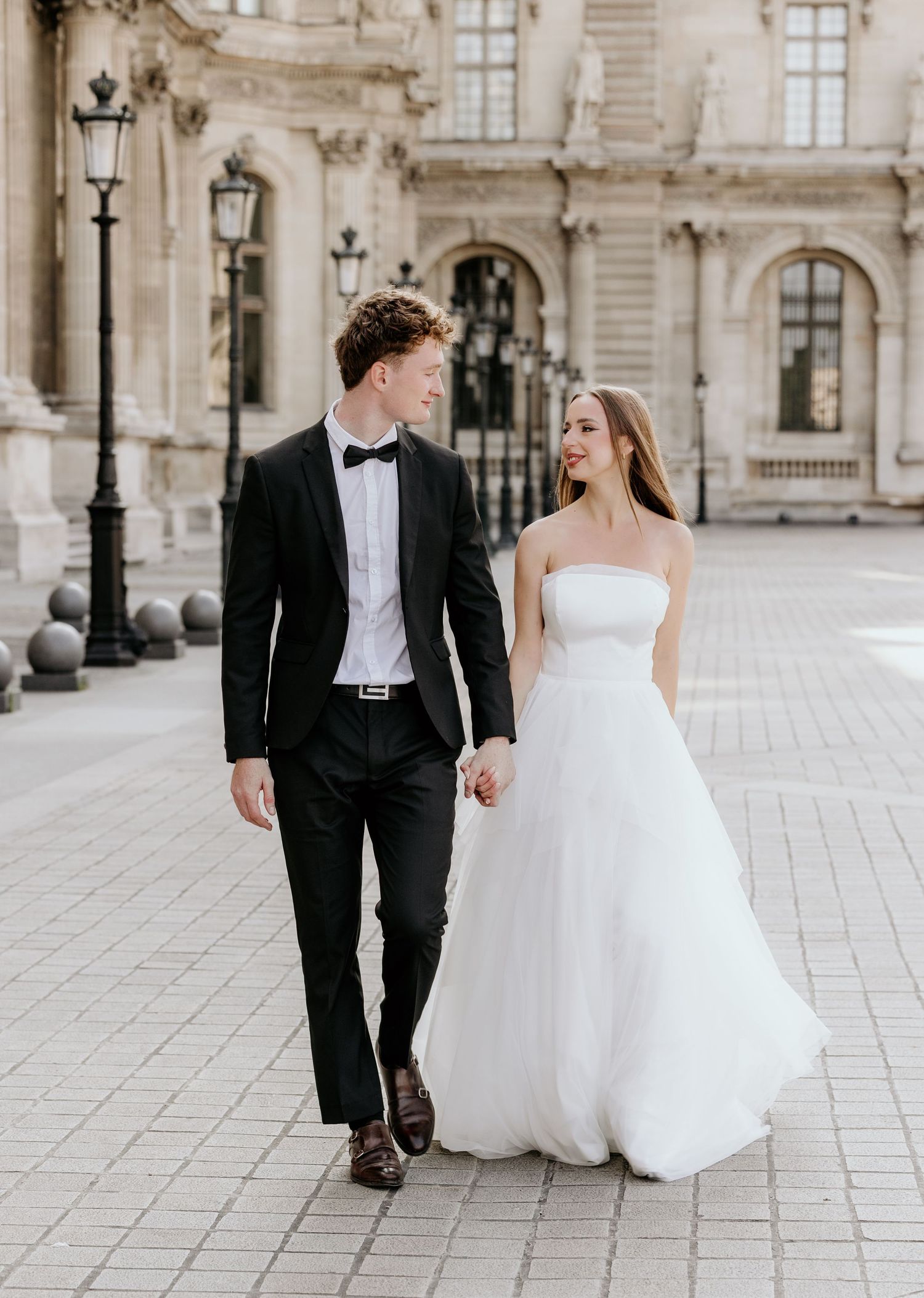 A bride and groom share joyful moments walking together on a grand stone courtyard with elegant architecture in the background.