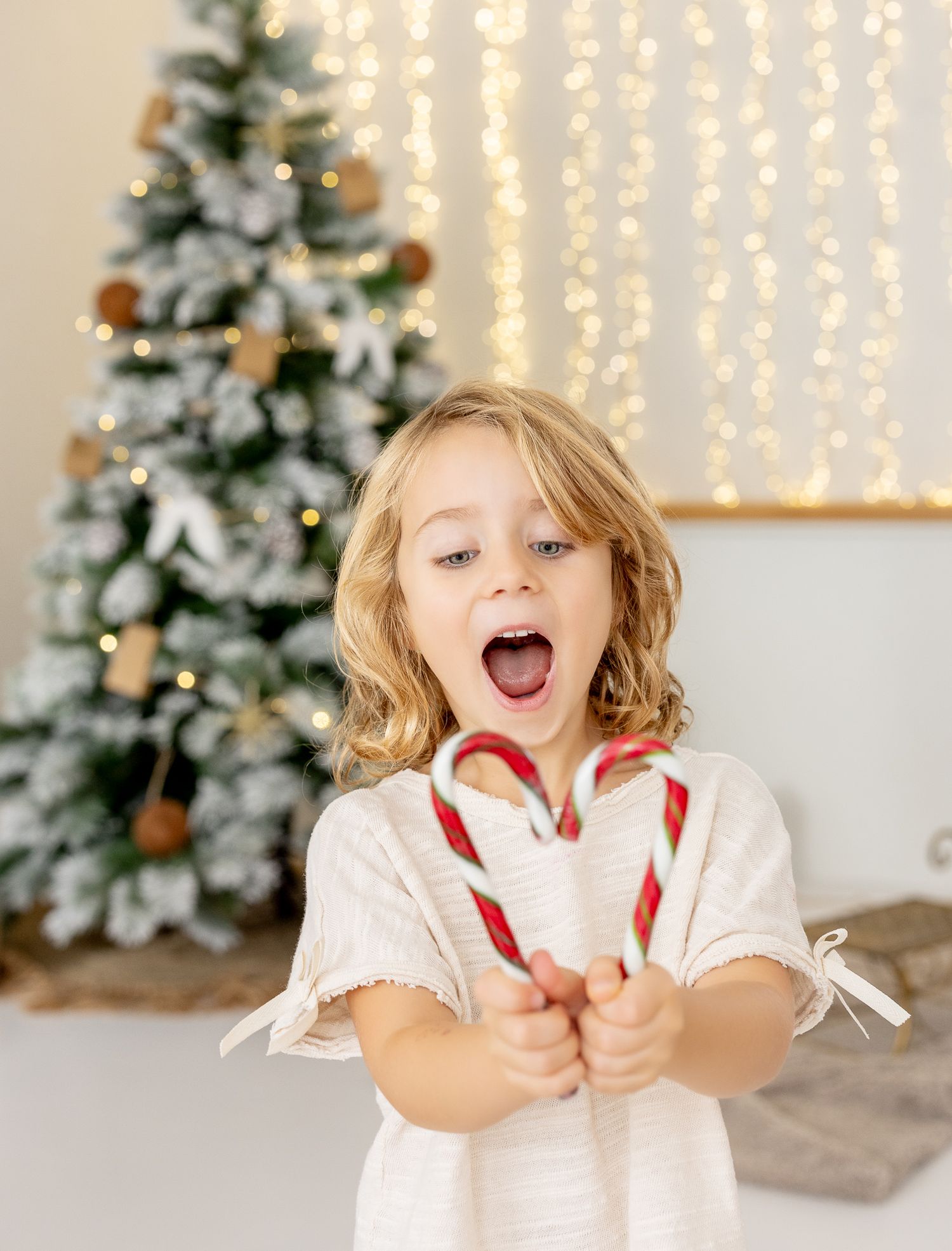 Child in white dress holding candy canes stands excitedly in front of decorated Christmas tree with twinkle lights.