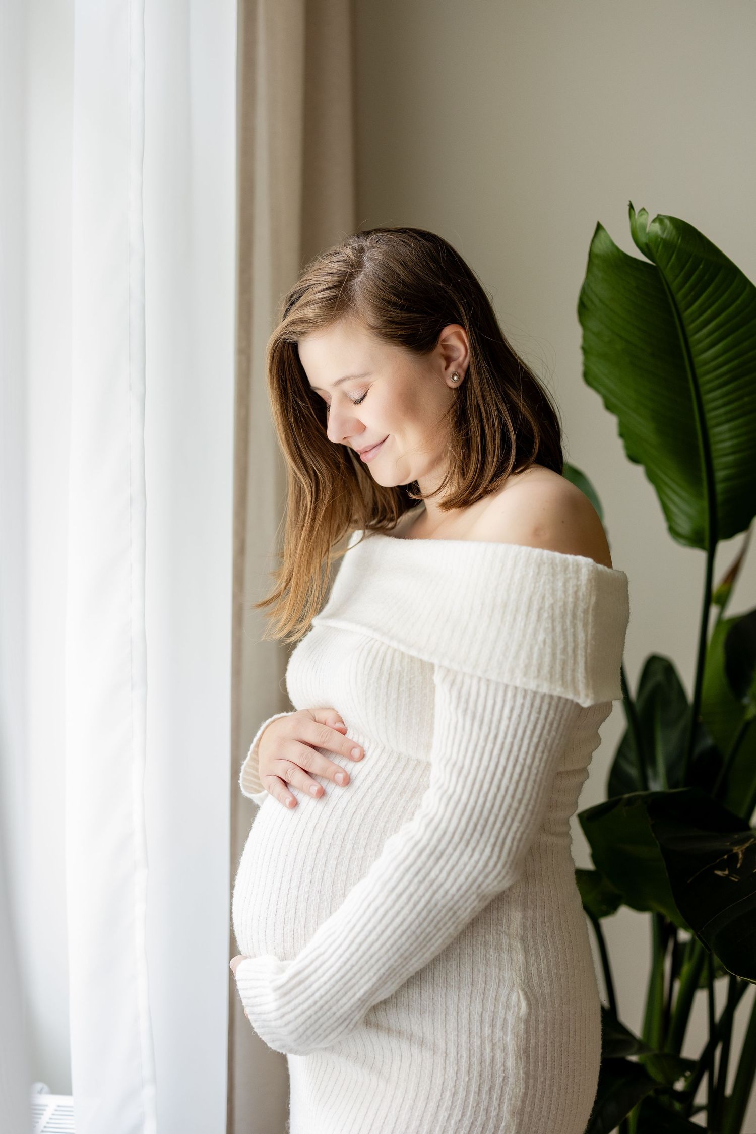 Pregnant woman in white off-shoulder knit dress cradling her baby bump near a bright window with tropical plant.