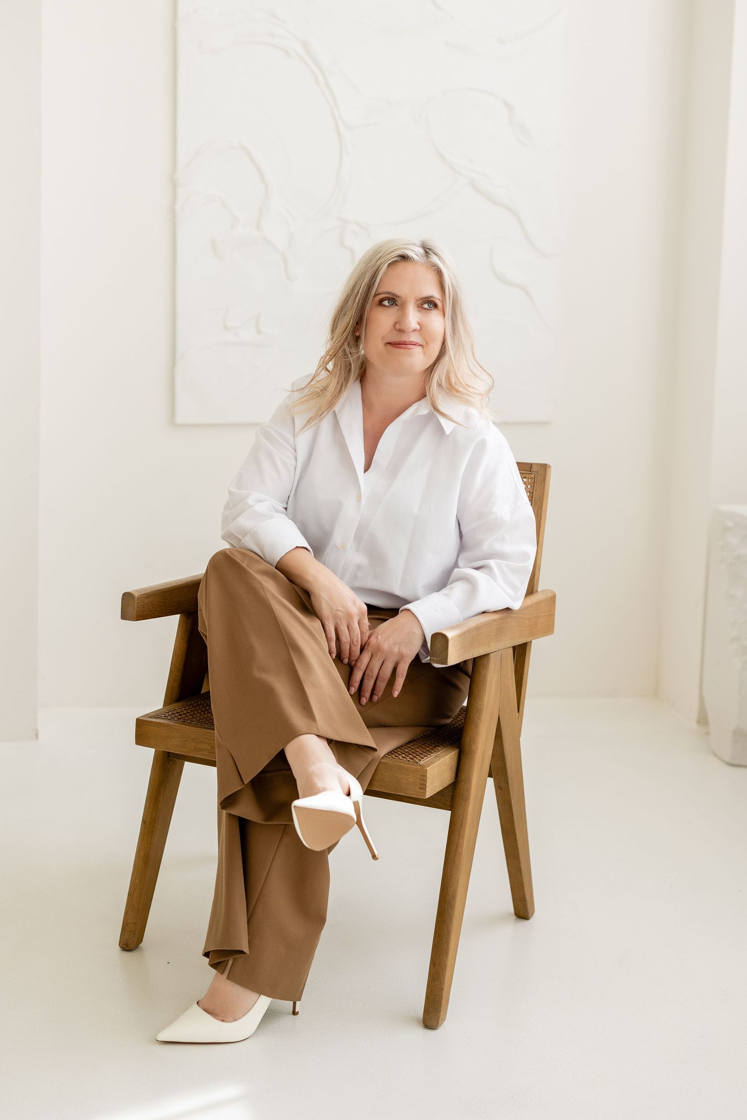 A person in a crisp white shirt and tan trousers sits casually in a wooden chair against a minimalist white background.
