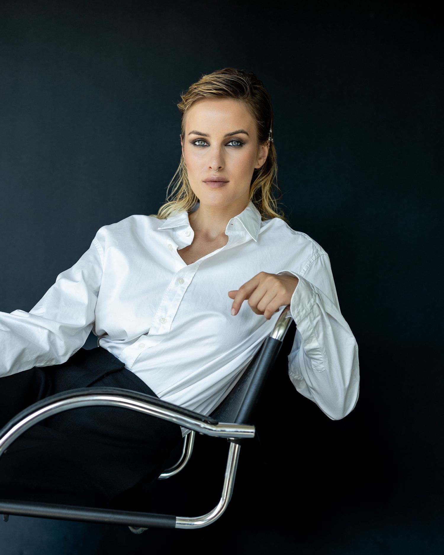 Blonde woman in white blouse seated in modern chrome chair against dark background, confident professional pose.