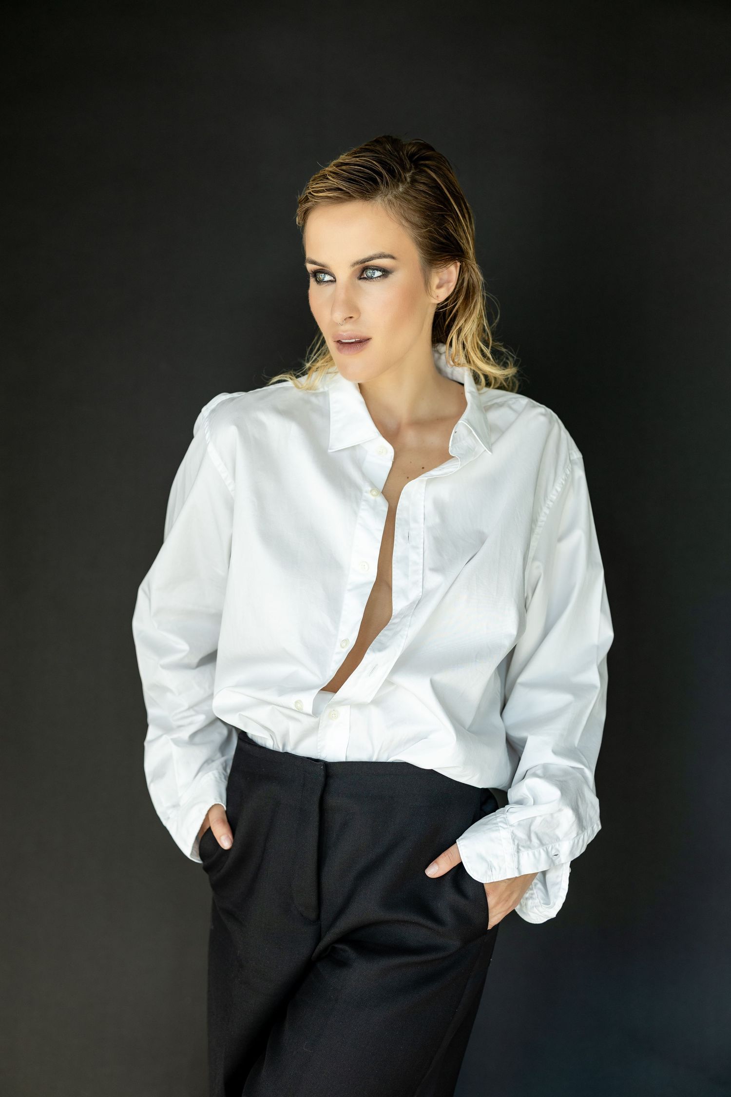 Woman in white button-up shirt and black trousers posing confidently against dark background.