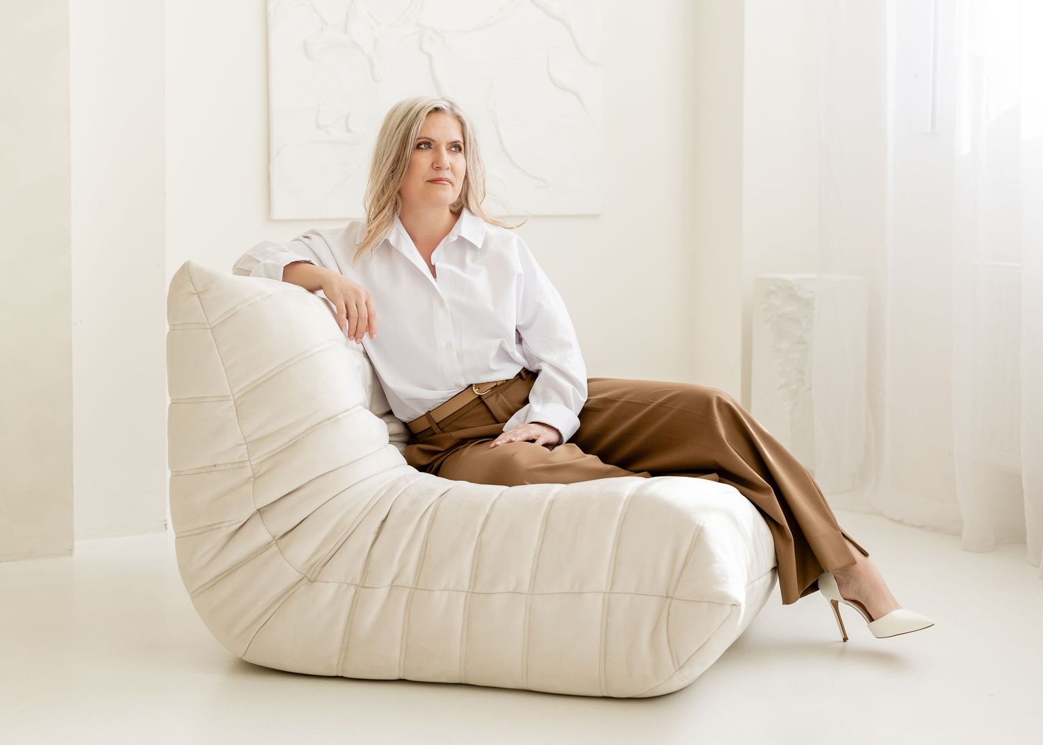 A person in a white shirt and brown pants relaxes on a plush cream-colored bean bag chair in a bright room.