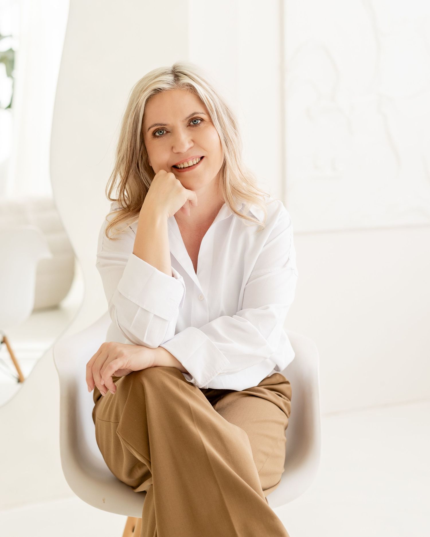 A person in a white button-down shirt and tan pants sits thoughtfully in a bright, minimalist setting.