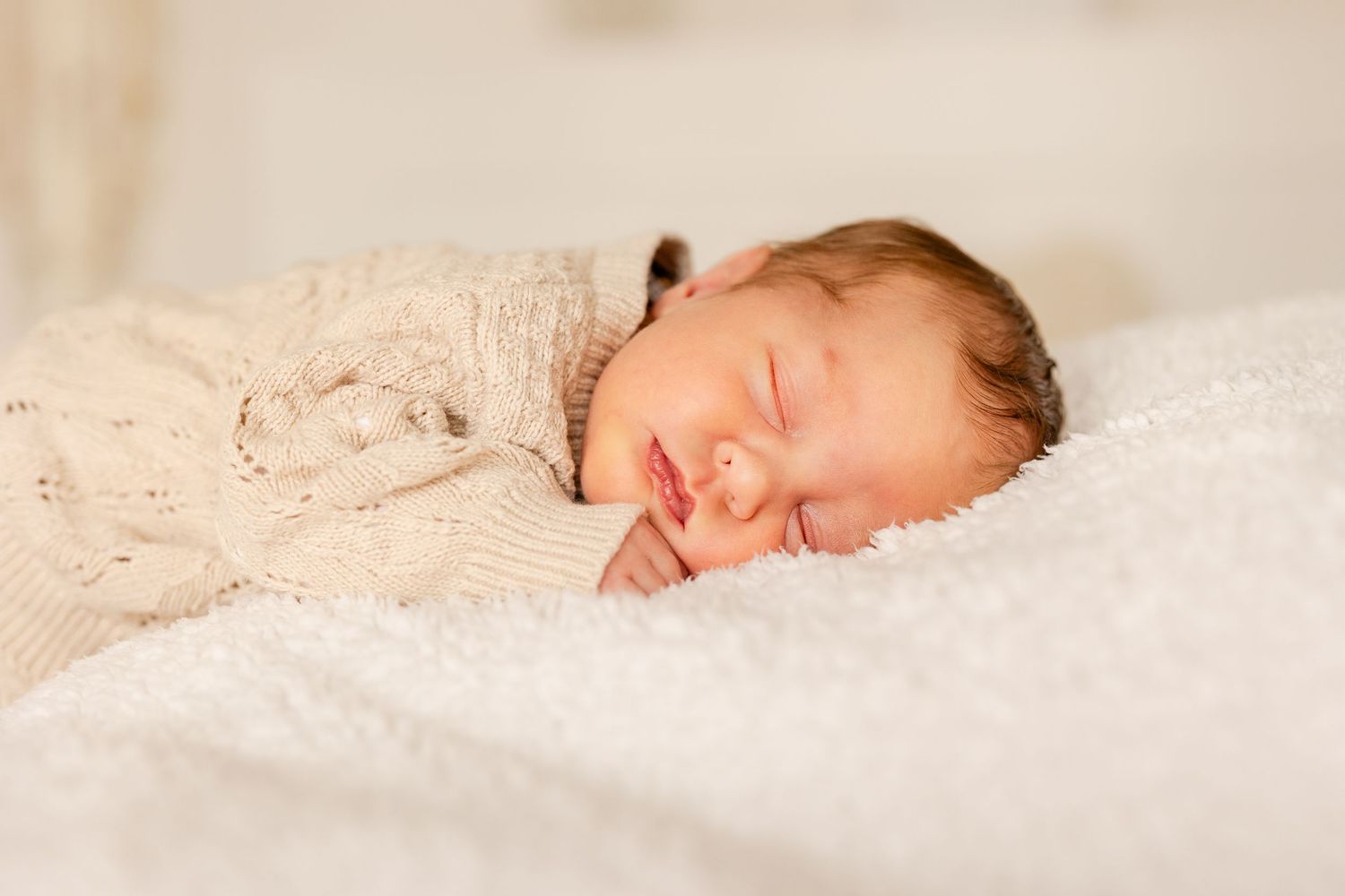 Peaceful newborn baby sleeping on white fluffy blanket wearing a cozy knit sweater.