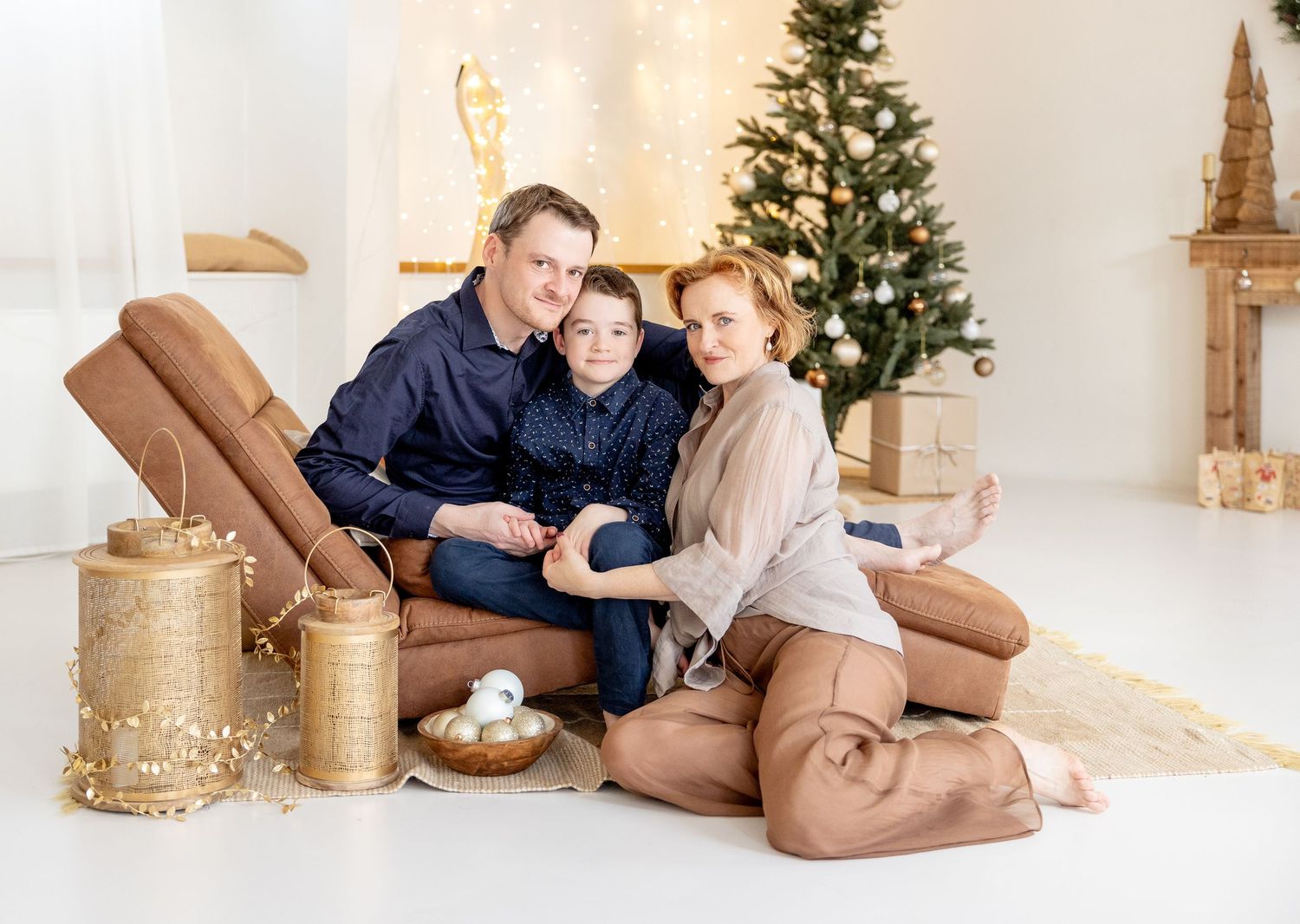 A family relaxes together on a beige couch beside a Christmas tree and decorative gold lanterns.