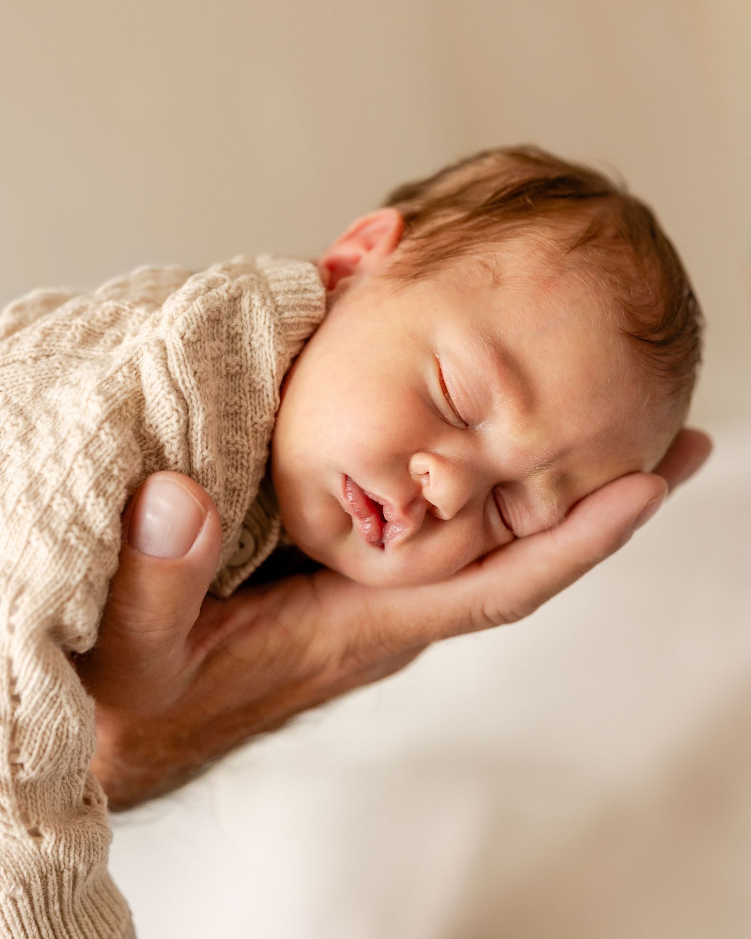 Newborn baby sleeping peacefully on an adult hand, wrapped in a cozy beige knit sweater.