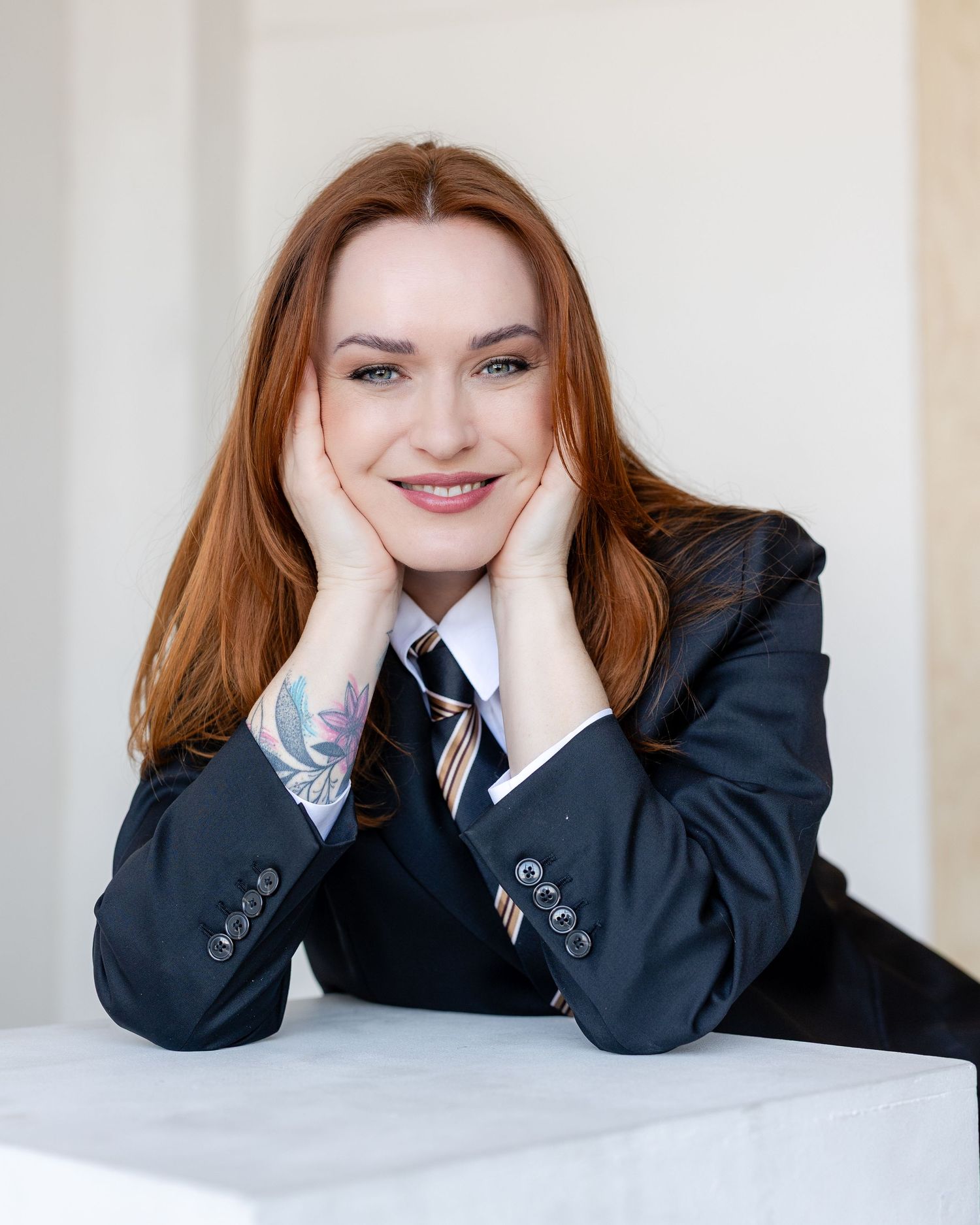 Smiling redhead woman in a dark blazer with striped tie, resting face on hands, posing for a professional portrait photo.