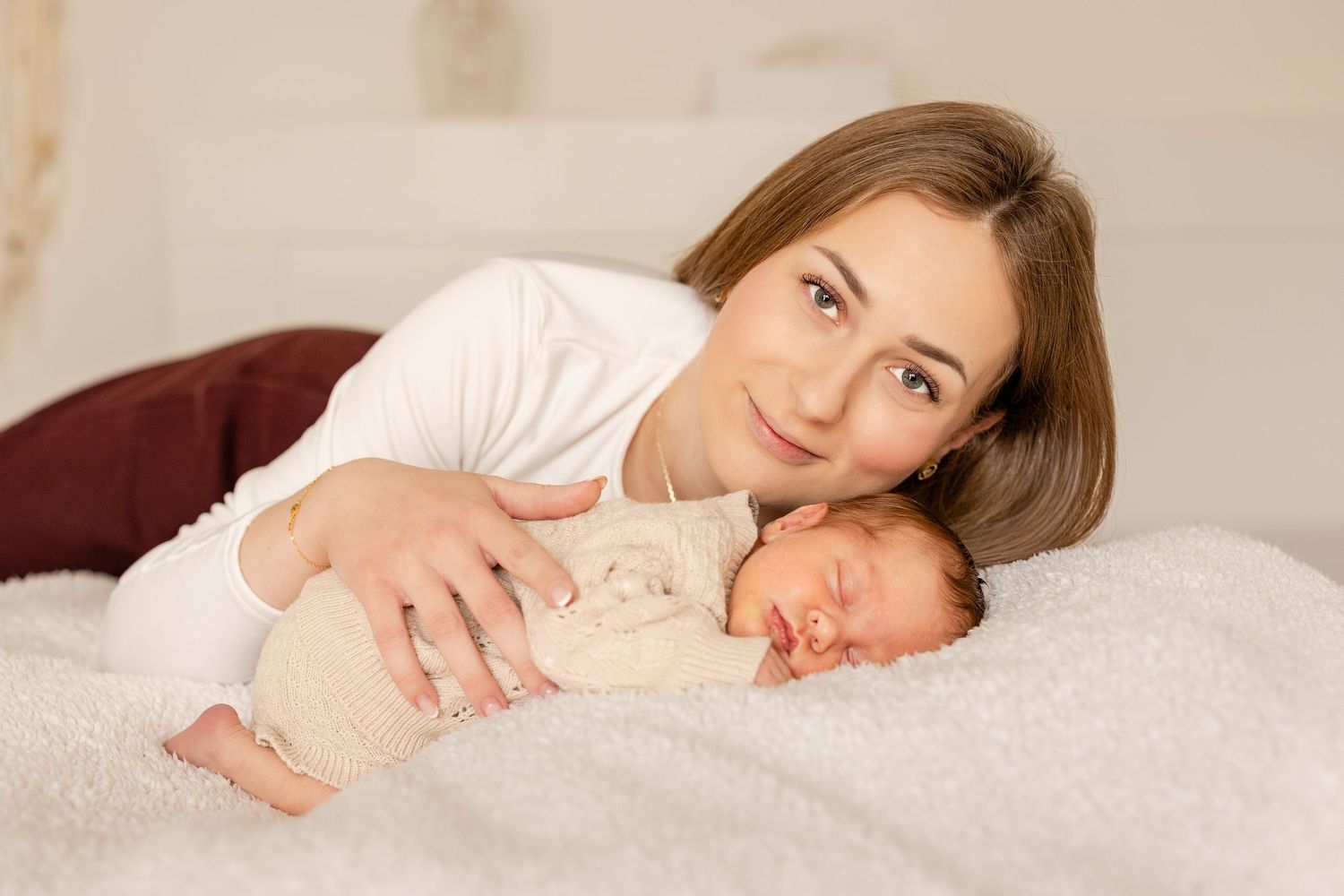 A young mother lies on a white bed smiling, gently holding her sleeping newborn baby dressed in a beige outfit.