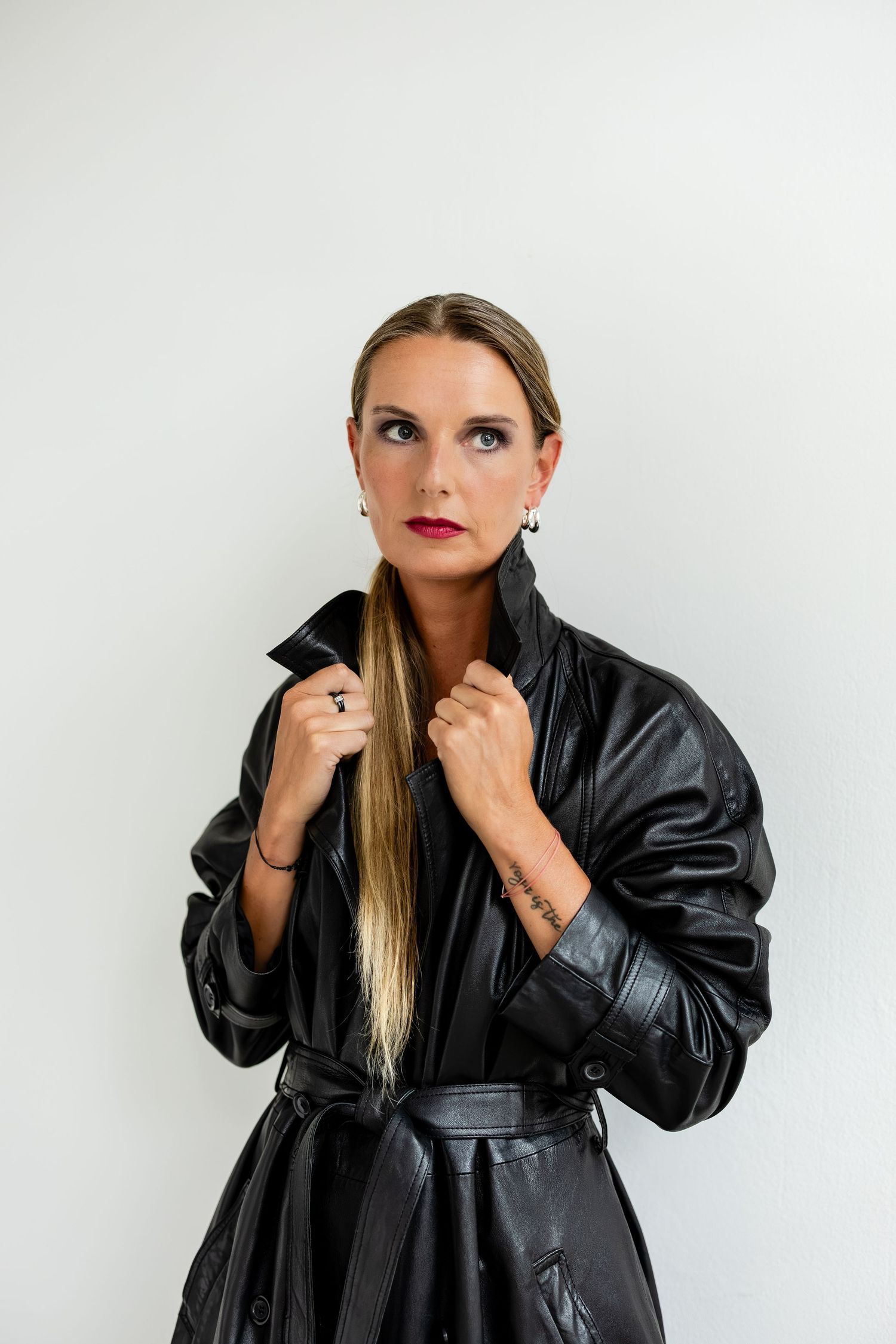 Woman in black leather trench coat with red lips and blonde hair posing against white background.